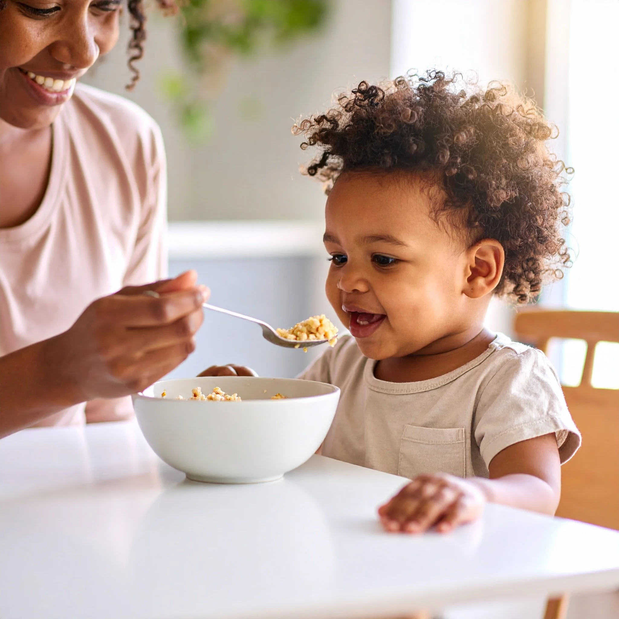 A mother feeding her smiling toddler at a table