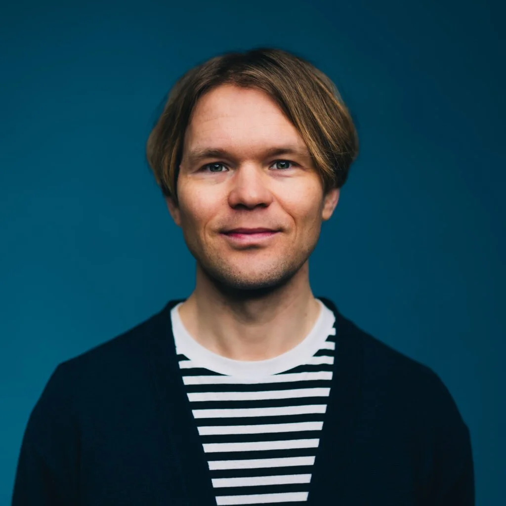 Portrait of a man with light brown hair, wearing a black cardigan over a black and white striped shirt, against a solid dark blue background.