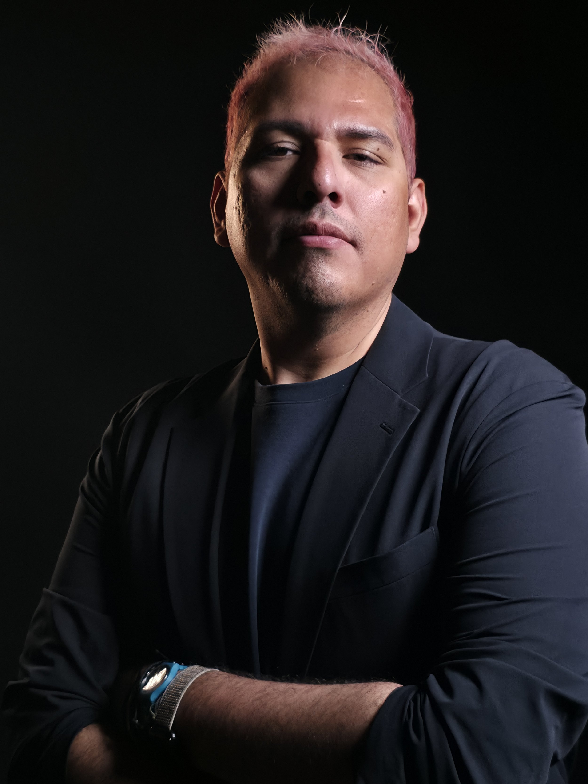A man with pink hair wearing a black blazer and t-shirt, crossing his arms and looking confidently at the camera against a dark background.