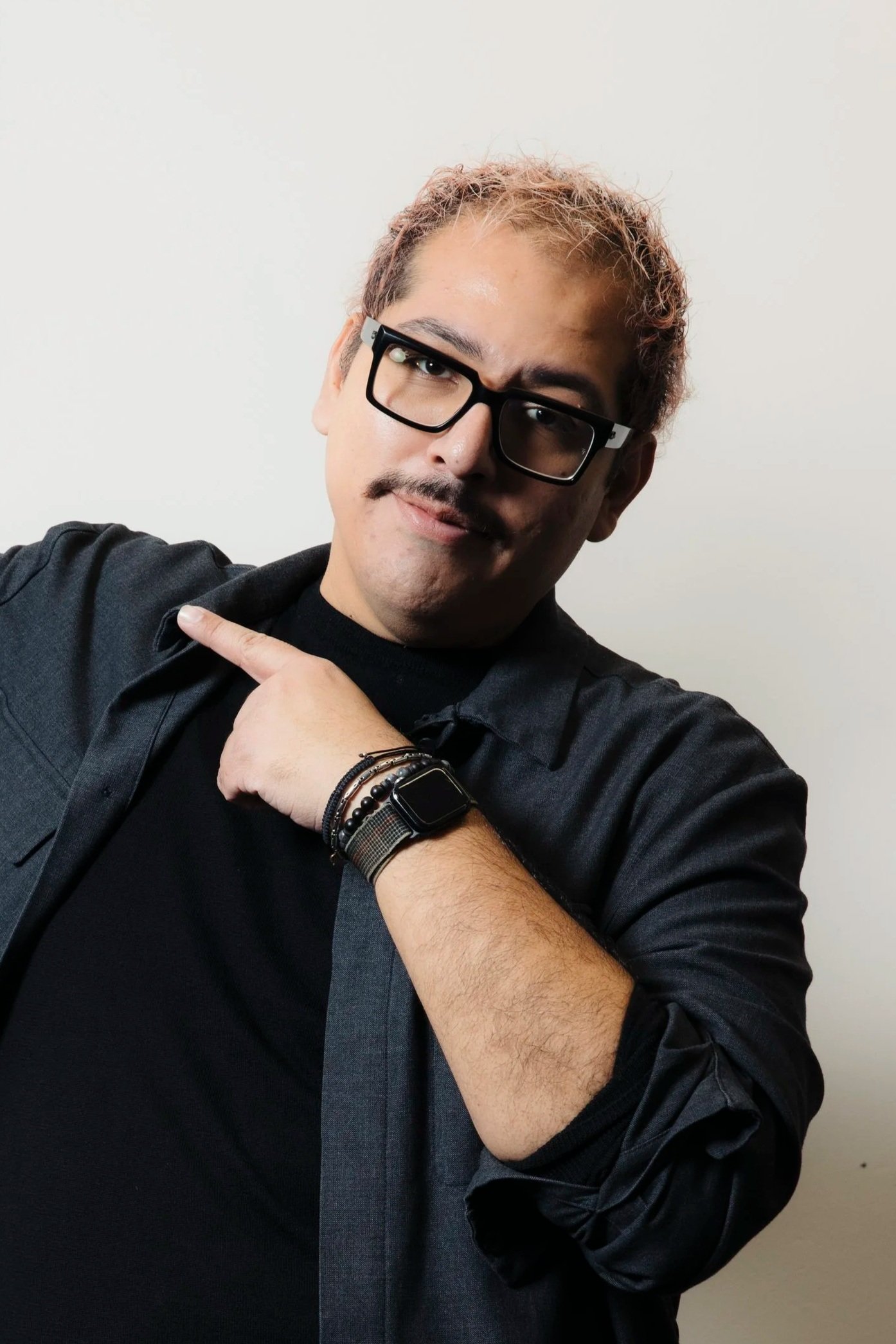 A man with glasses, a mustache, and short hair, wearing a black shirt and a gray jacket, pointing to his left with a confident expression, against a plain white background.