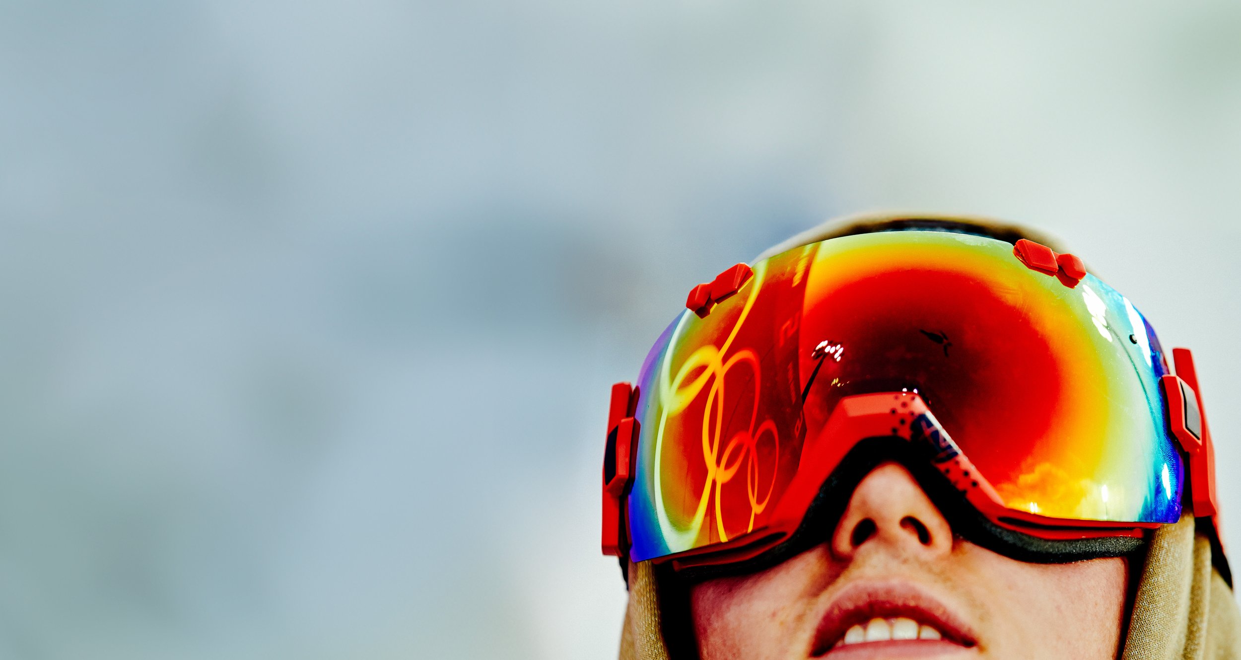 Close-up of a person wearing goggles with a rainbow-colored reflective lens, partially visible facial features, and a cloudy sky in the background.
