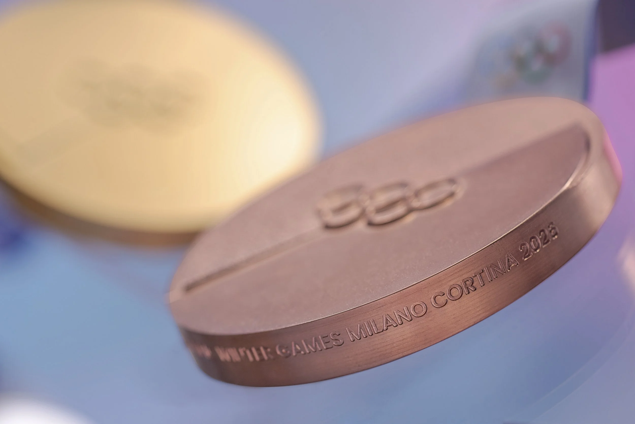 Close-up of a bronze-colored medal with the Olympic rings and text 'GAMES MILANO CORTINA 2026' engraved on its edge.