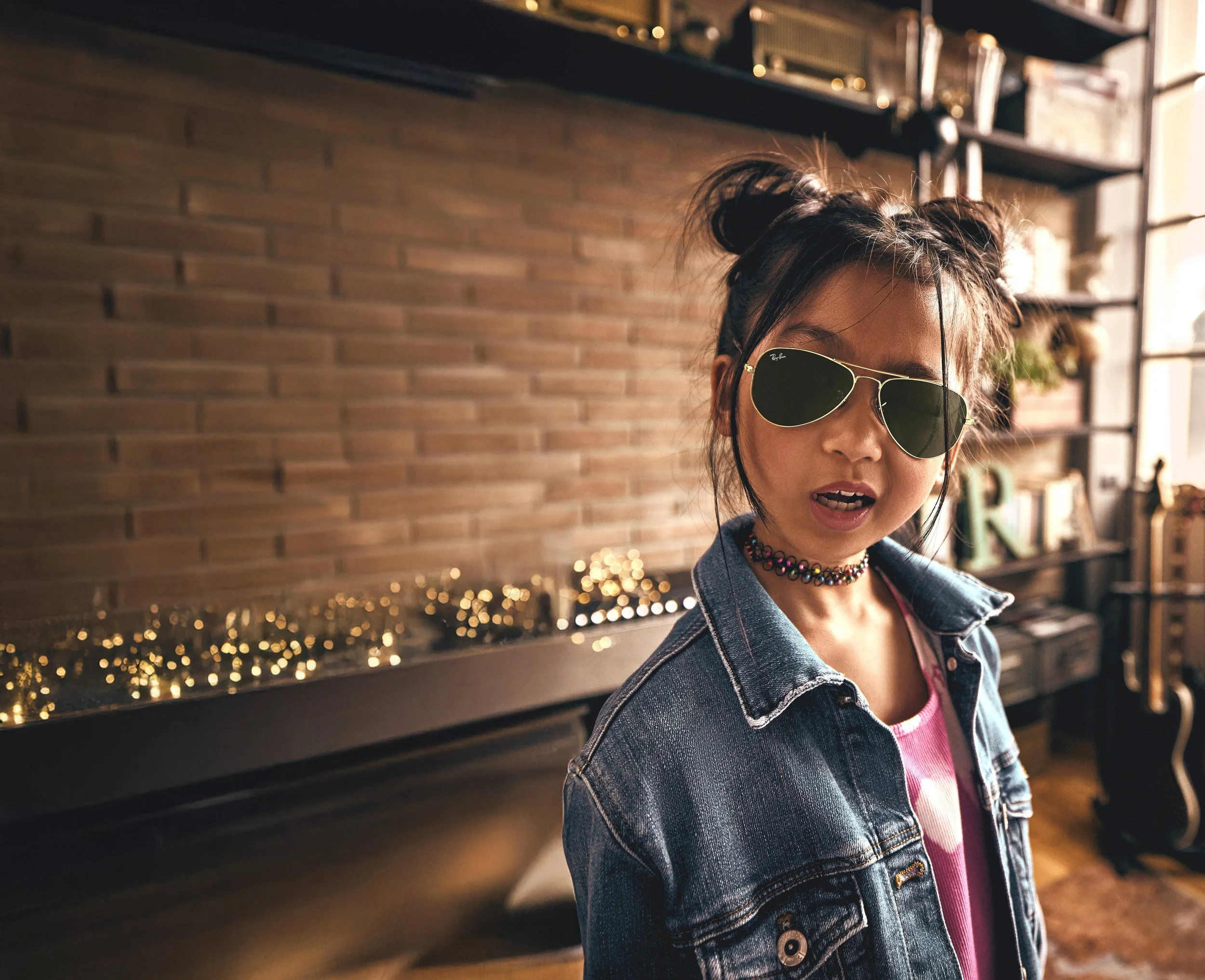 A girl with dark hair styled in buns wearing aviator sunglasses, a pink shirt, a denim jacket, and a choker necklace, standing indoors with a brick wall and shelves in the background.