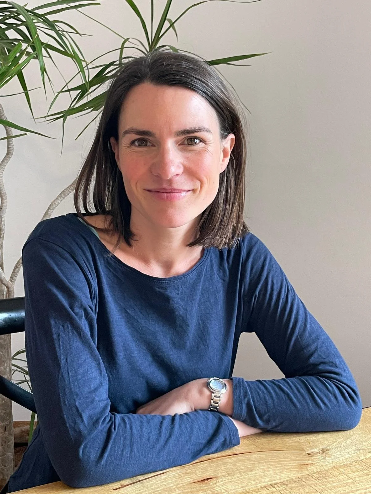 Une femme souriante assise à une table en bois, portant un sweat bleu, avec un bracelet en acier à la montre, derrière elle un grand plante verte.