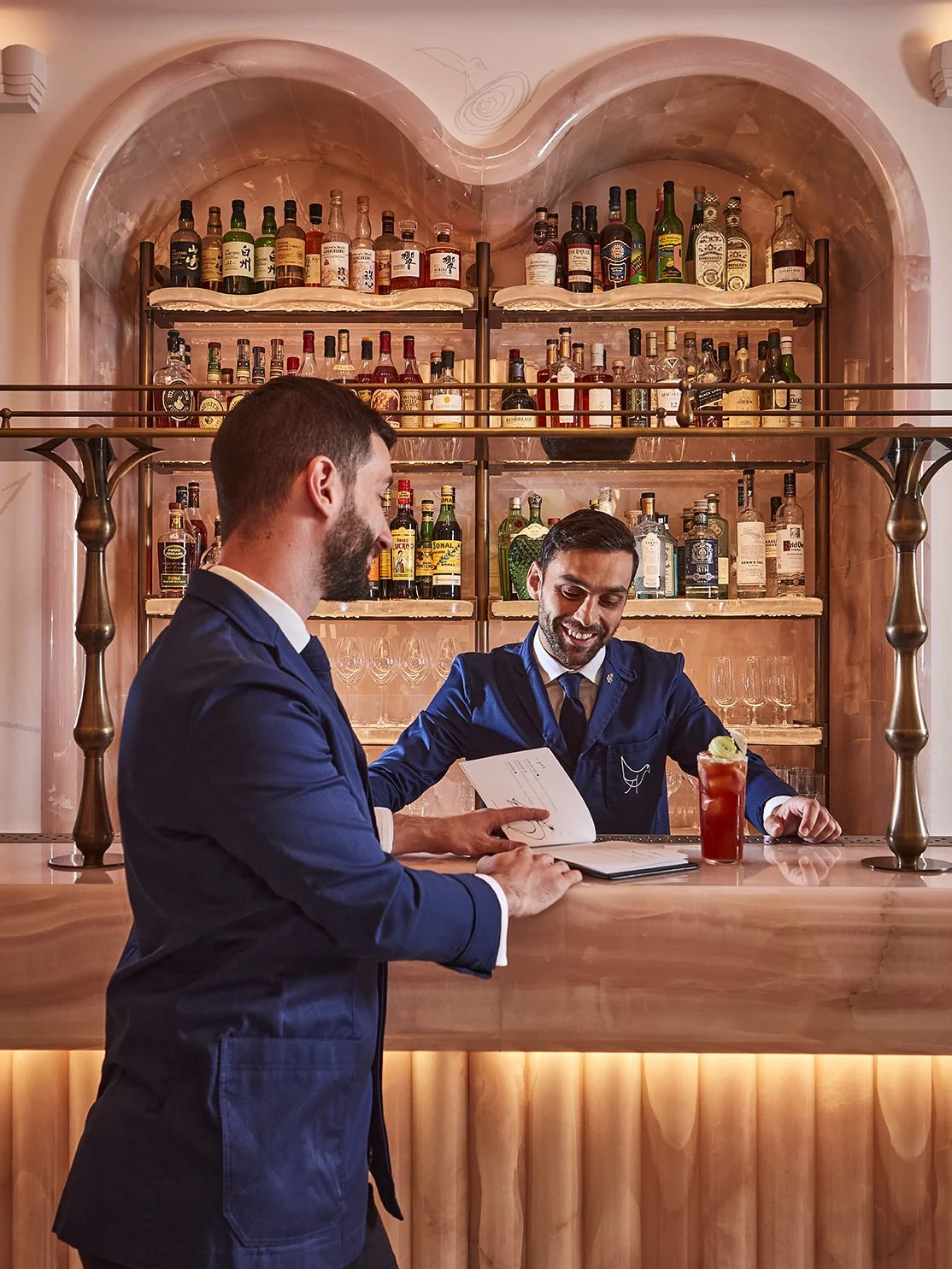 Two men in suits at a bar, one holding a menu, the other smiling with a drink, behind a wooden bar with shelves of liquor bottles.
