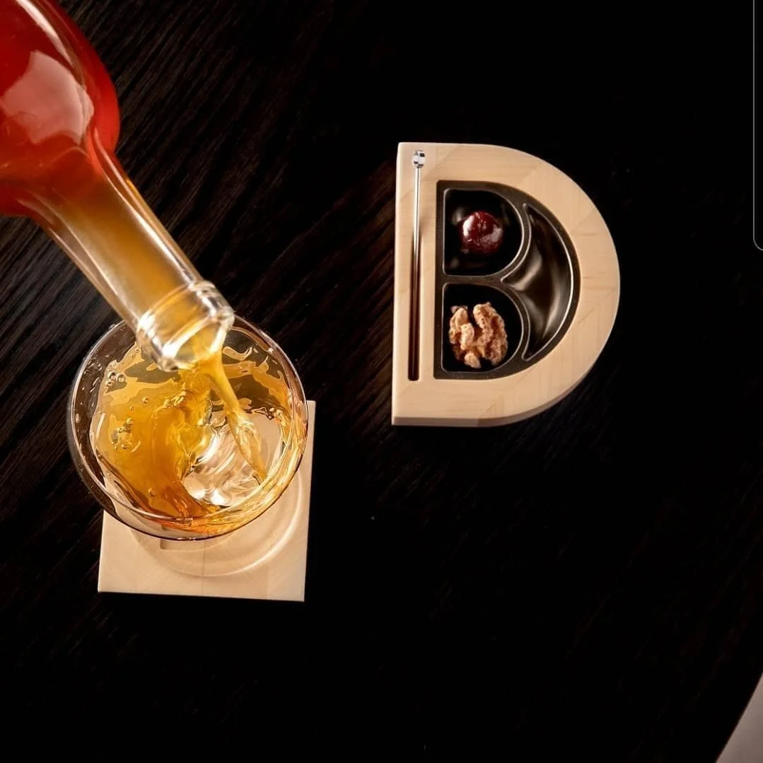 A glass being filled with a golden-colored beverage from a bottle, placed on a coaster on a dark wooden table; a small wooden divided tray with compartments holding a cherry and some snack bits is nearby.