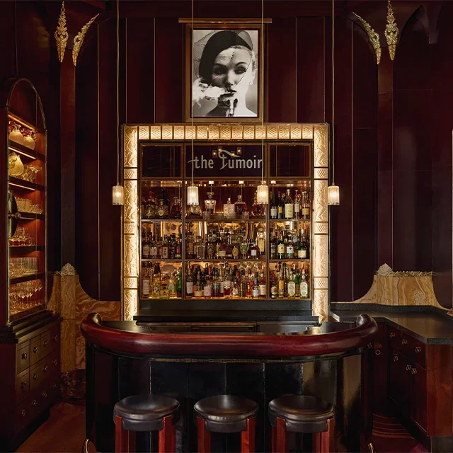 The image features a luxurious bar interior with a dark wood theme, illuminated by warm lighting. Behind the bar, there are shelves stocked with various bottles of liquor and bar accessories. Above the bar, a framed black-and-white portrait of a woman is displayed, and a sign reads "the Tûmour." The bar has a curved red countertop with three black bar stools in front of it.