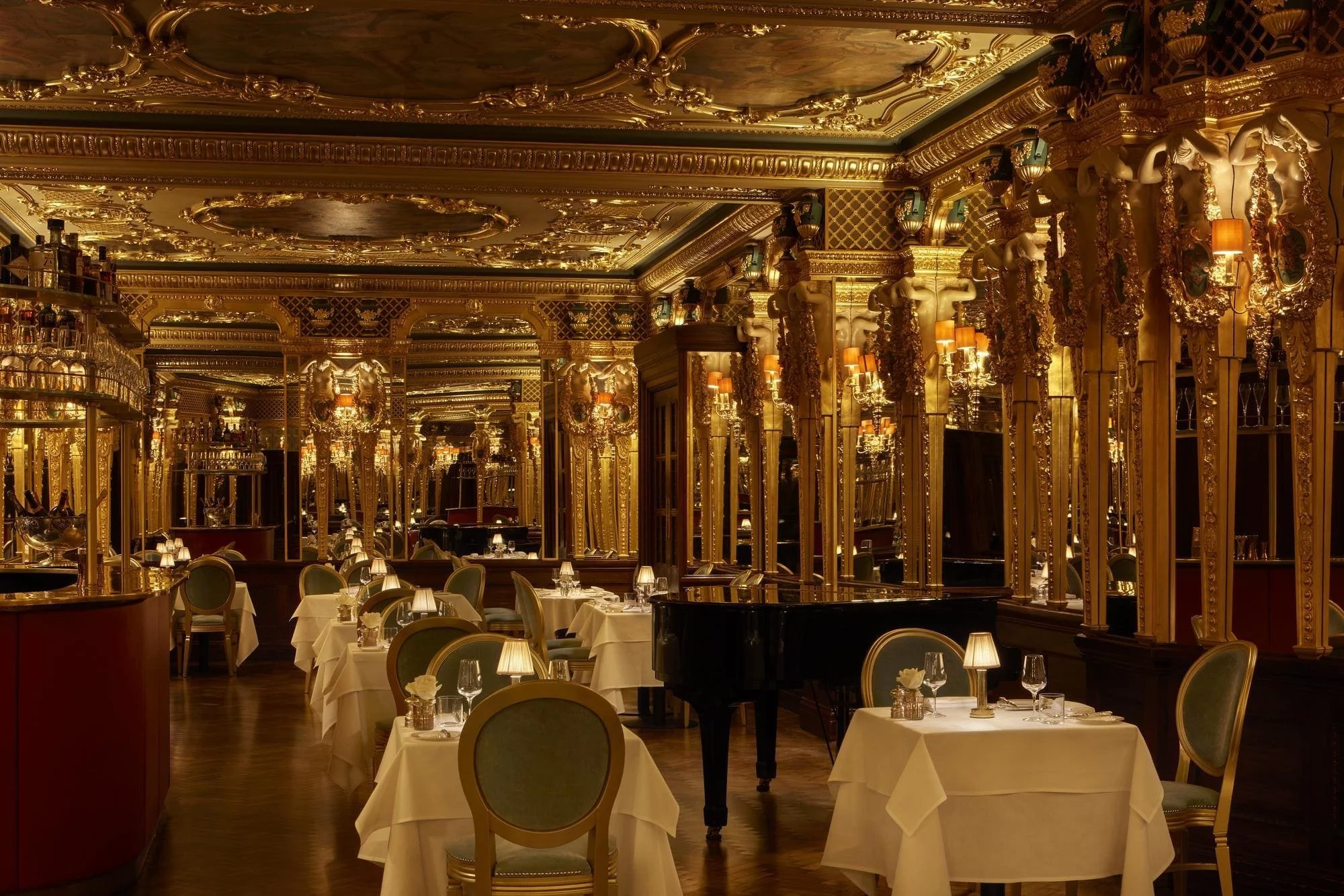 Luxurious restaurant interior with gold ornate decor, mirrors, chandeliers, tables with white tablecloths, and a black grand piano.