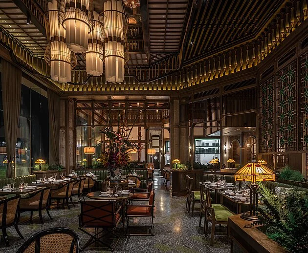 Interior of an elegant restaurant with dim lighting, large hanging lamps, a central floral arrangement, and neatly set tables with green and brown chairs.