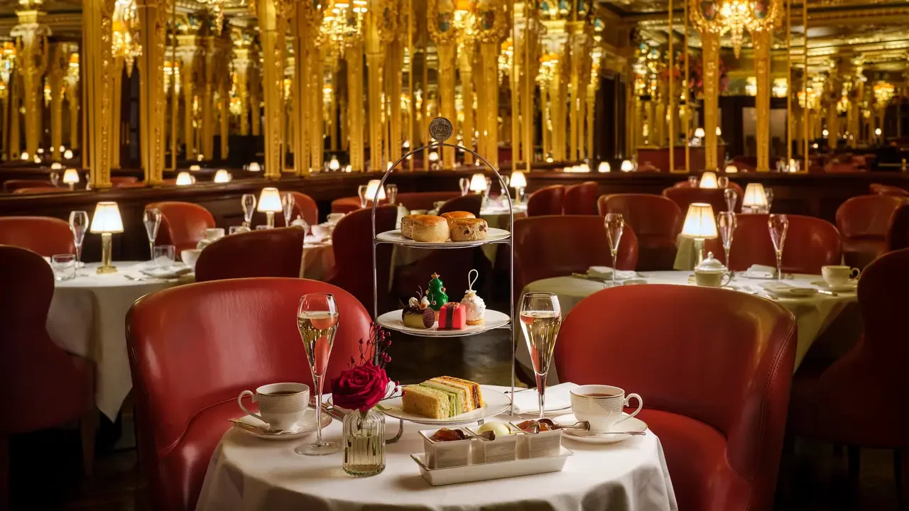 Elegant restaurant table set with white tablecloths, cups, glasses of champagne, and a flower vase, with a three-tiered dessert stand holding scones, sweets, and sandwiches, and red leather chairs, reflecting a luxurious atmosphere.