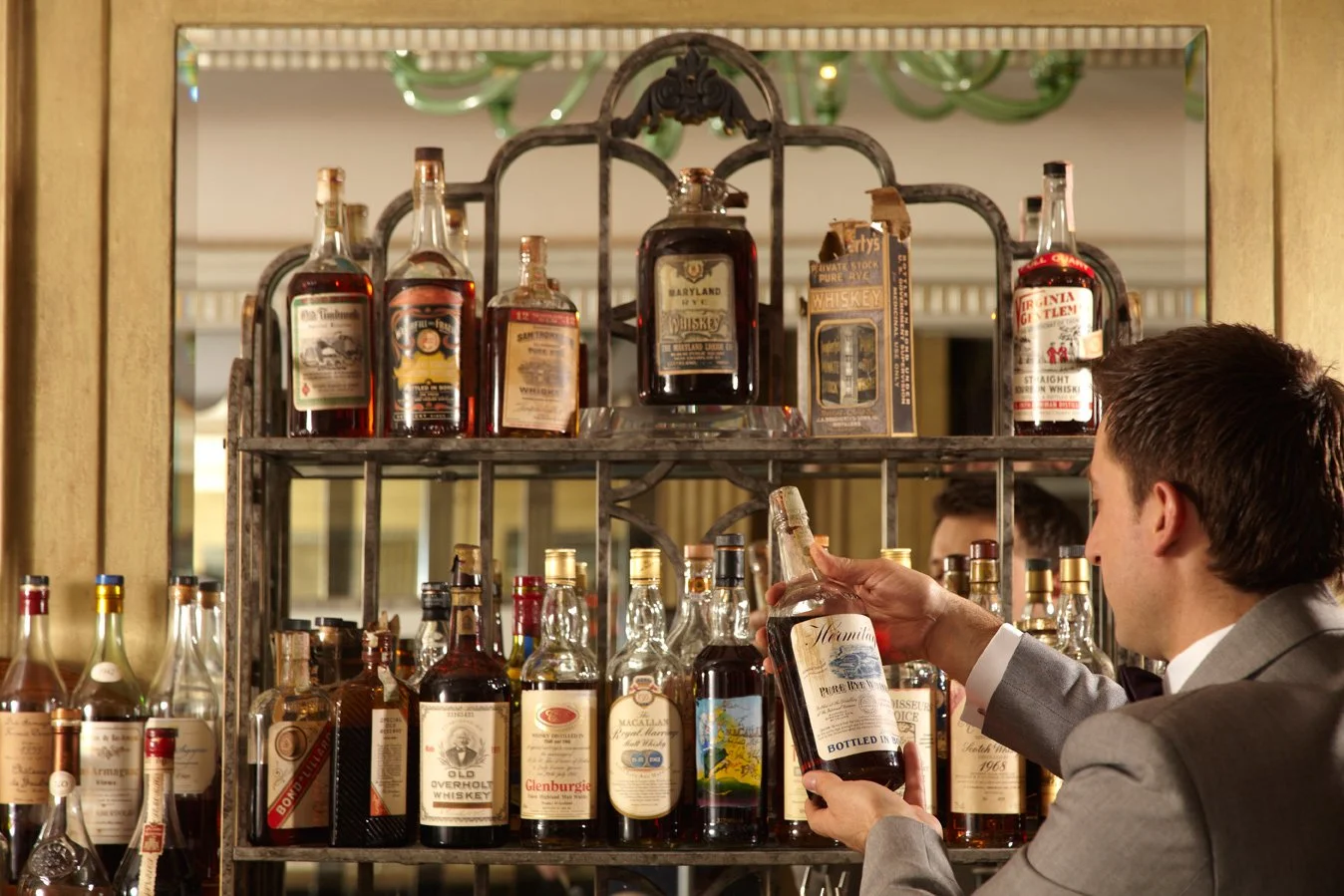 A bartender in a red tuxedo jacket holding a silver tray with a pink cocktail in a coupe glass and a glass of reddish drink garnished with a lemon slice.