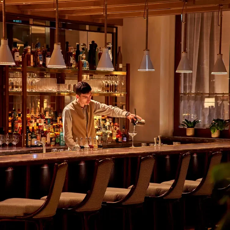 A bartender pouring a drink at a bar with liquor bottles and glasses behind him, and pendant lights hanging from the ceiling.