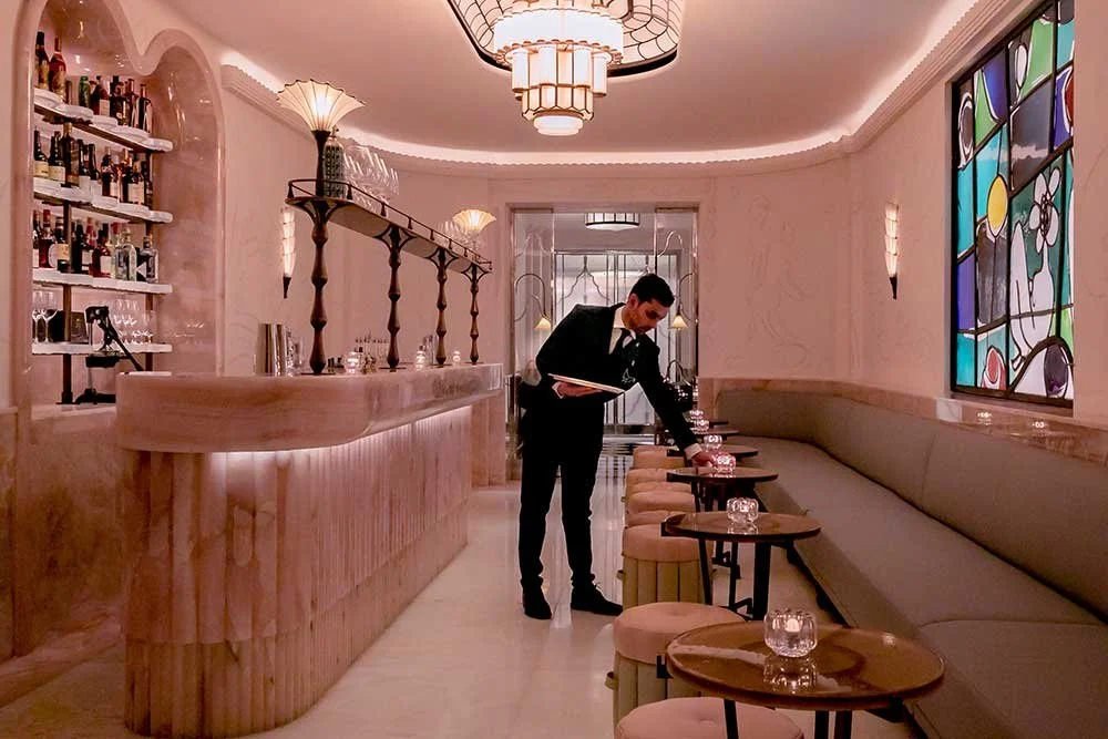 A waiter in a suit and tie adjusting a small table with a candle on it in an elegant restaurant or lounge with a contemporary design.