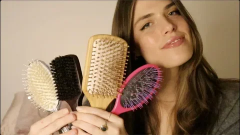 A young woman holding four different hair brushes, smiling at the camera.
