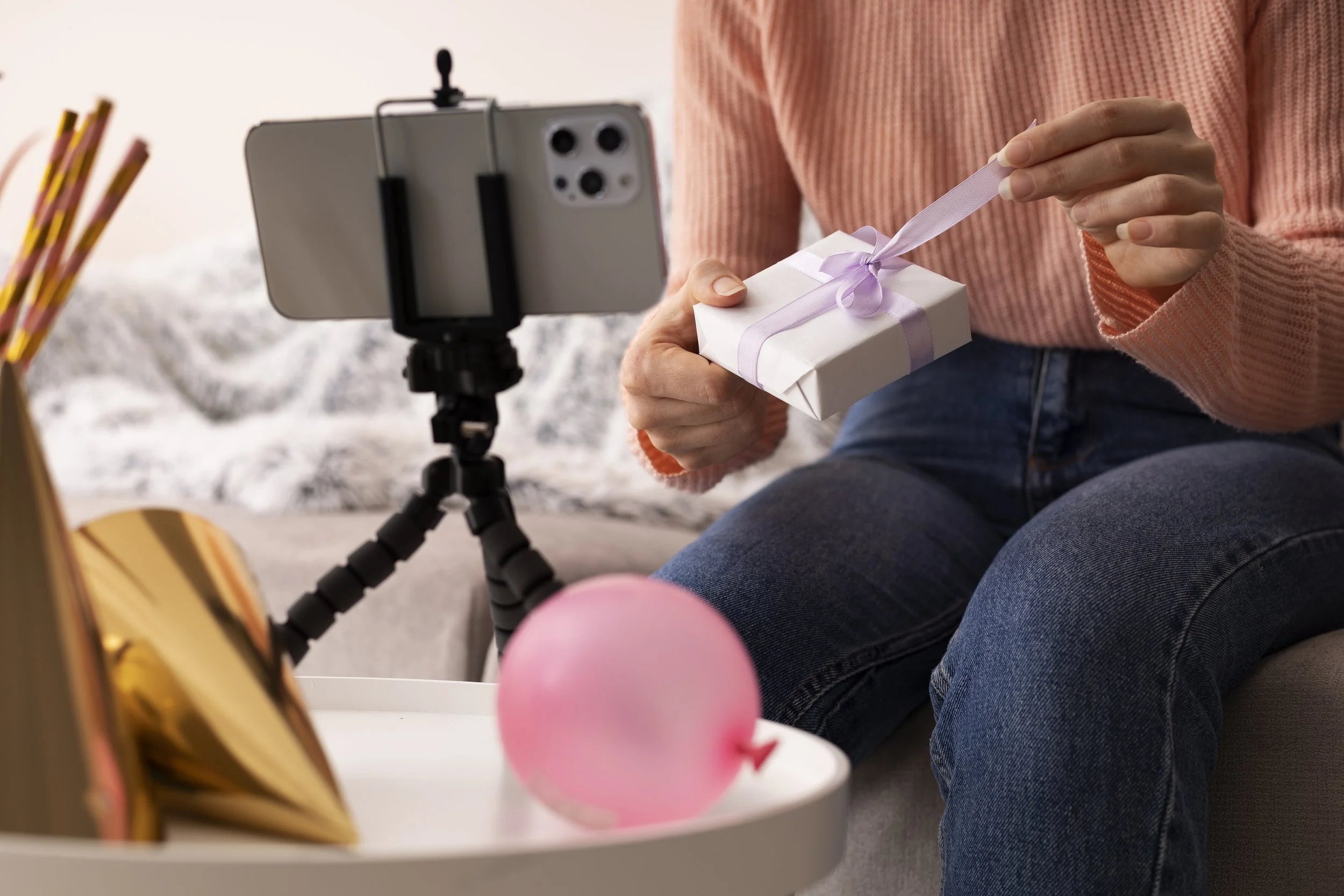 Person wrapping a small gift with white paper and a purple ribbon while filming with a smartphone on a tripod.