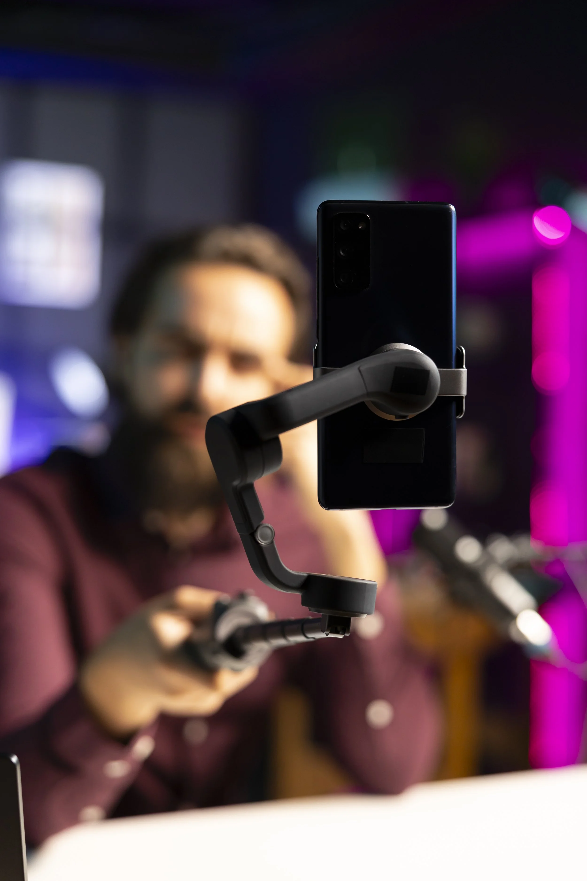 A man with glasses and a beard is holding a smartphone mounted on a stabilizer, taking a selfie or recording video. The background features colorful, blurred neon lights.