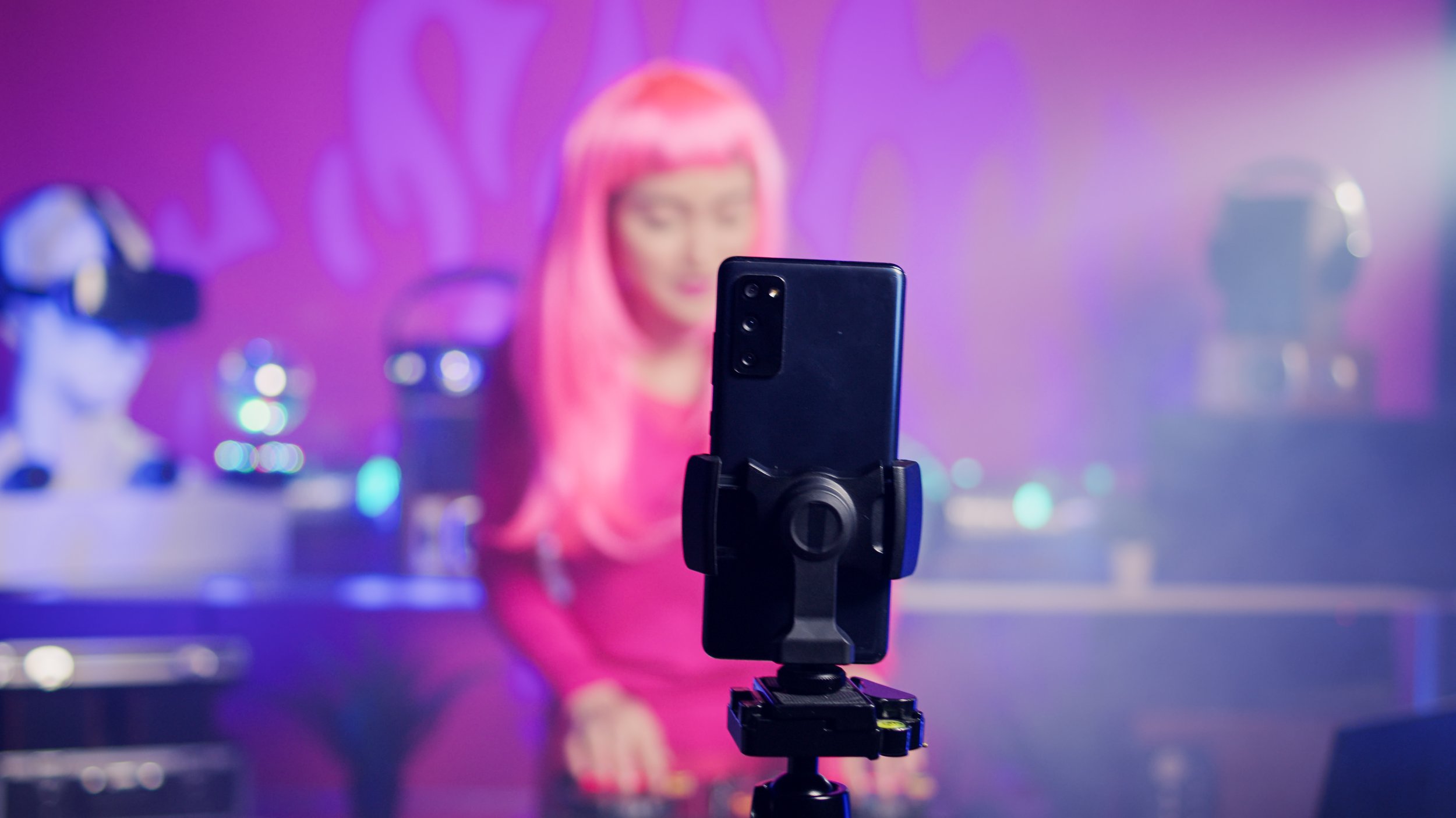 Smartphone mounted on a tripod in front of a woman with pink hair in a colorful, neon-lit setting.