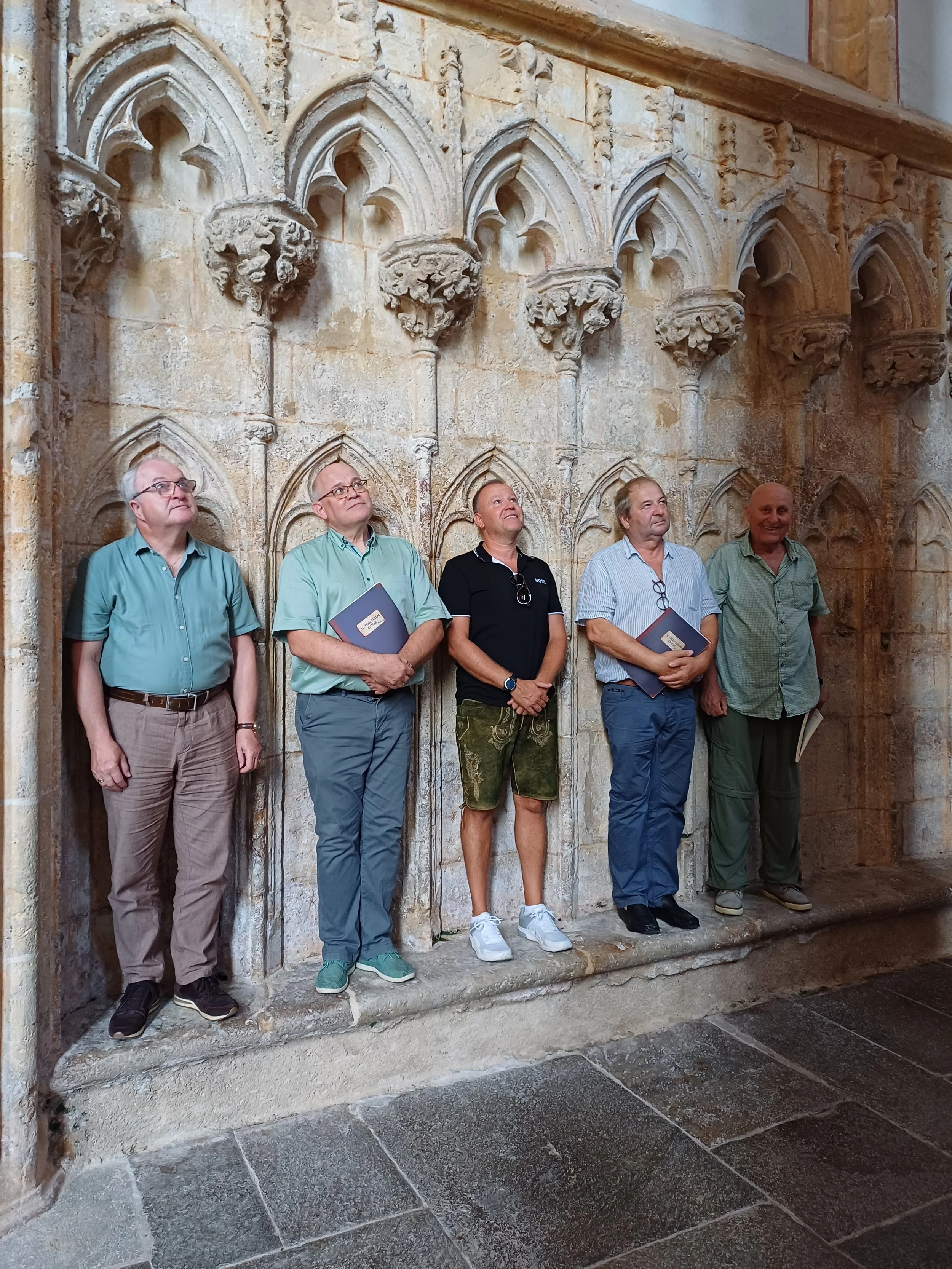Fünf Männer stehen an einer historischen Wand mit gotischer Architektur und schauen nach oben.