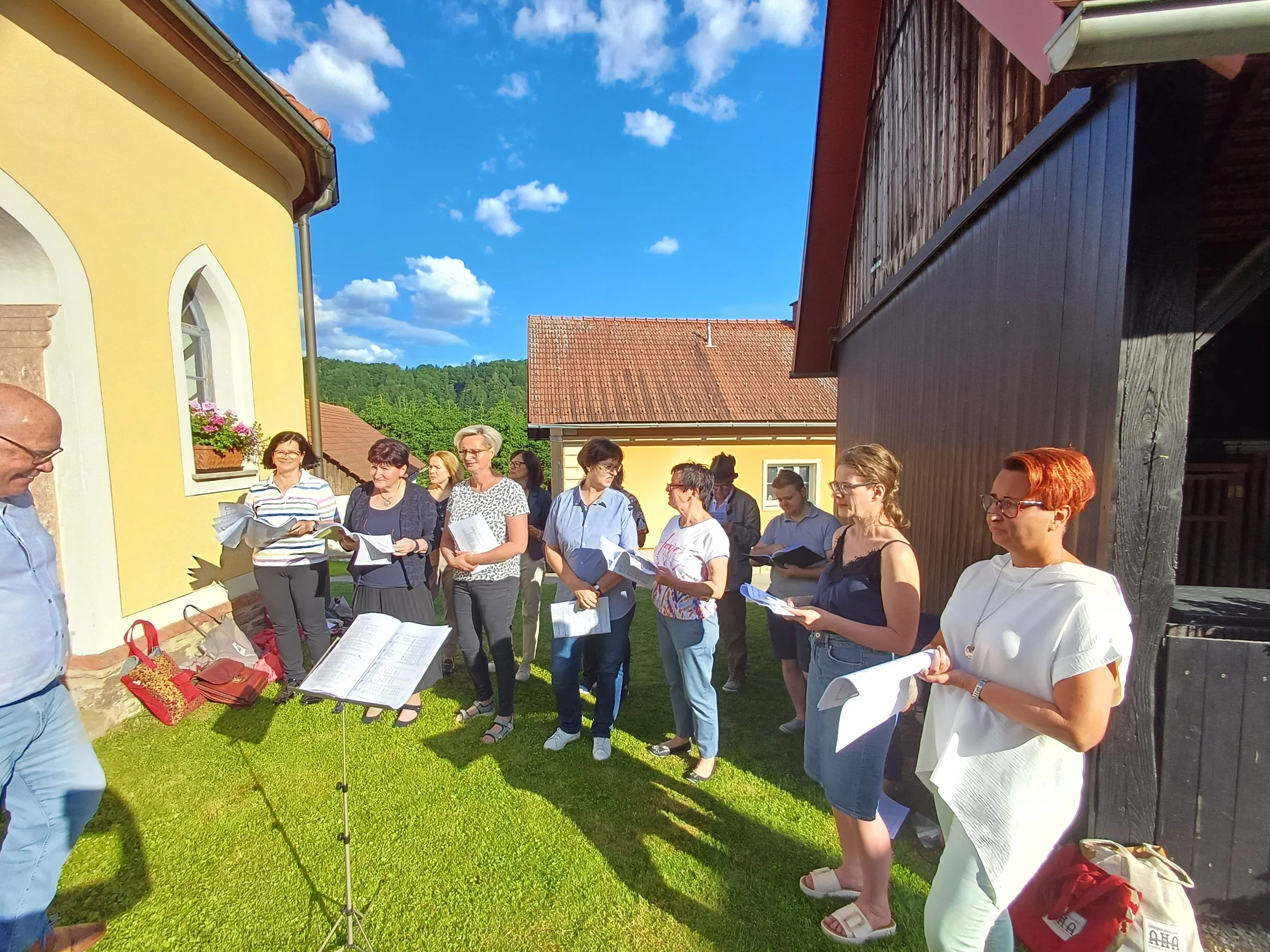 Eine Gruppe von Sänger:innen steht draußen im Sonnenschein vor einer gelben Kapellwand, hält Notenblätter in den Händen, während im Hintergrund grüne Hügel und ein blauer Himmel mit Wolken sichtbar sind.