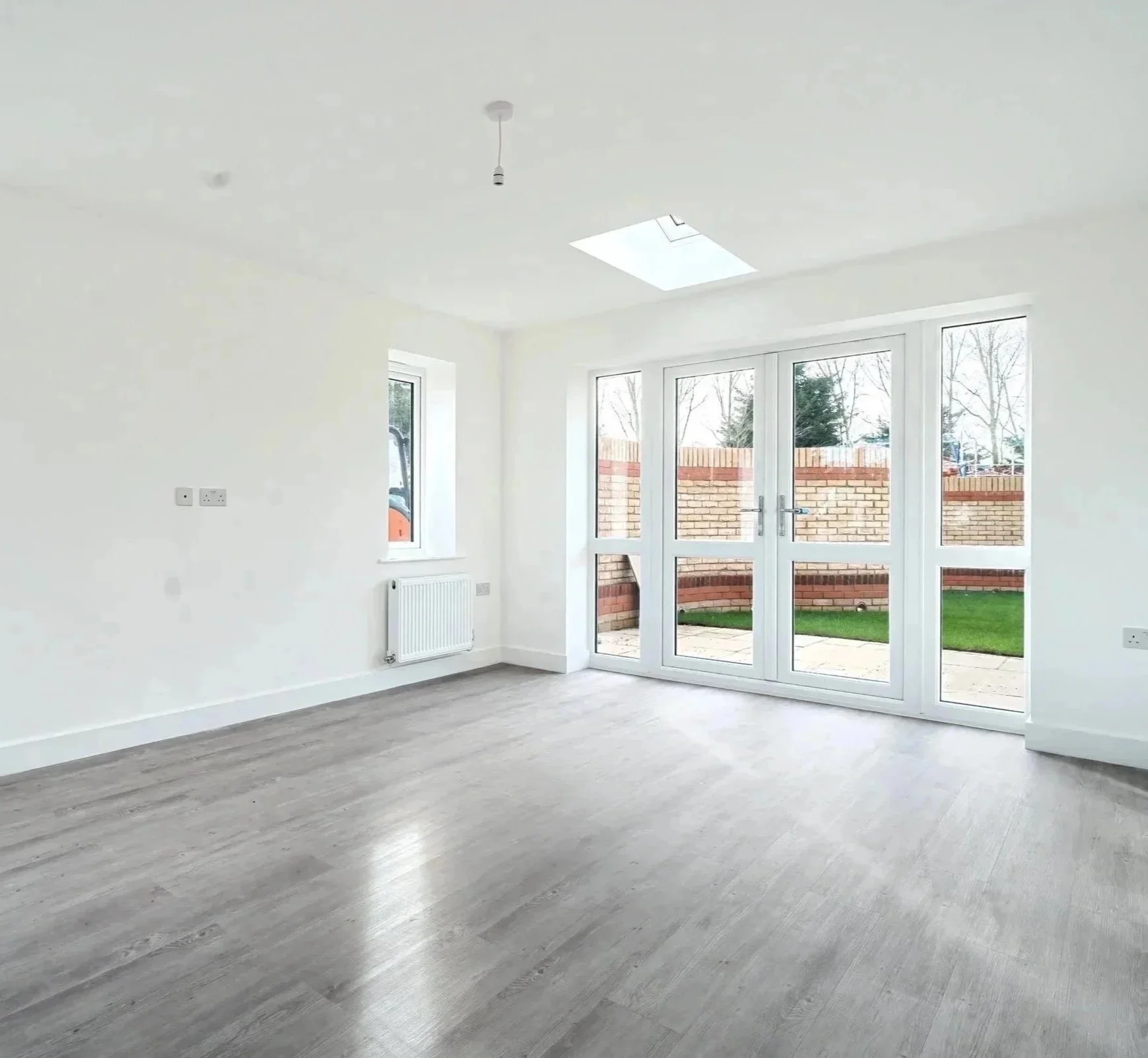 Empty living room with large glass doors leading outside, white walls, ceiling with skylight, gray wood flooring, and a radiator beneath a window.