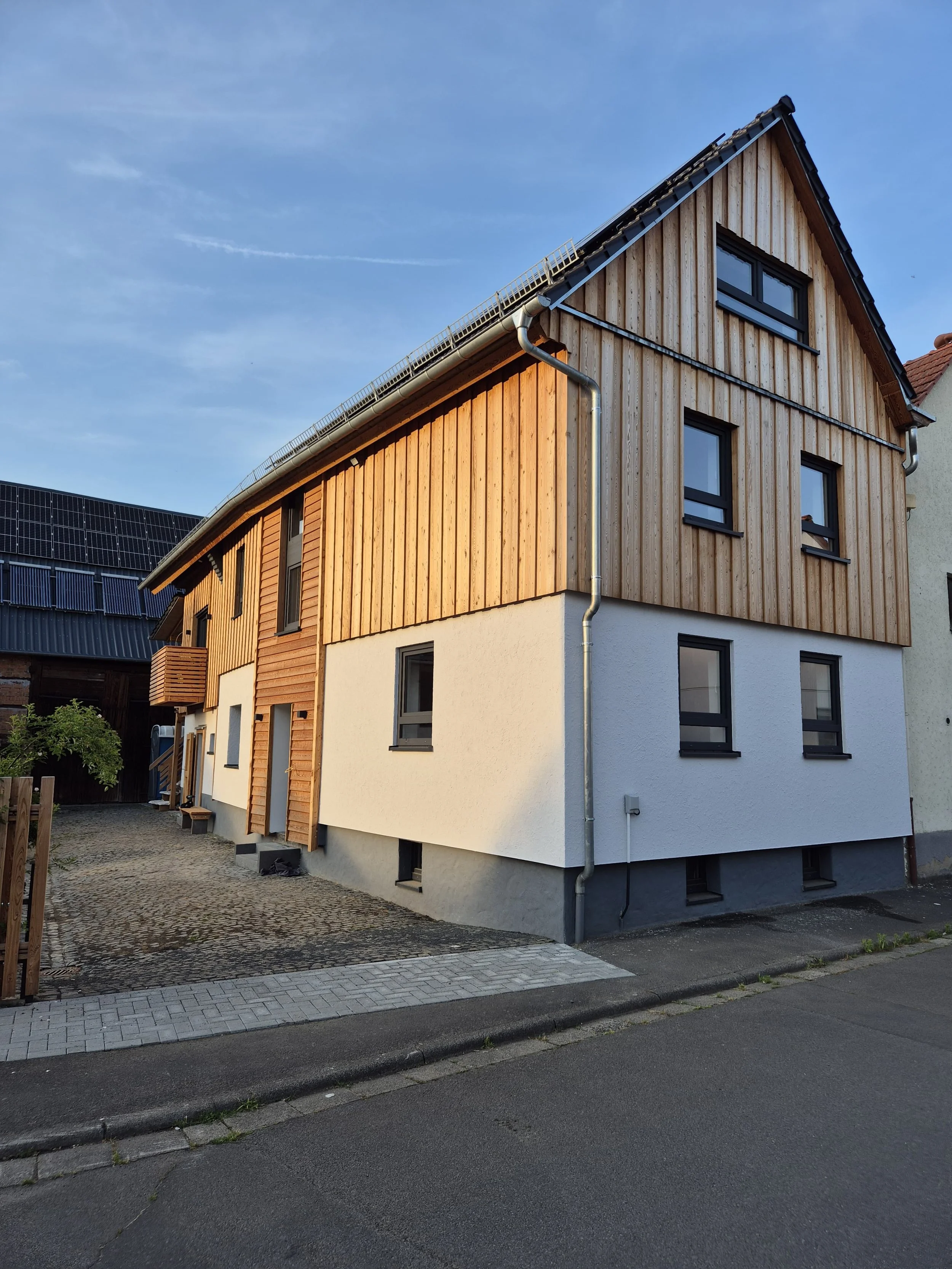 Mehrstöckiges Haus mit Holzfassade an der Ecke einer Straße, Sonnenlicht auf der Fassade, sonniger Himmel, stromführende Kabel am Himmel.
