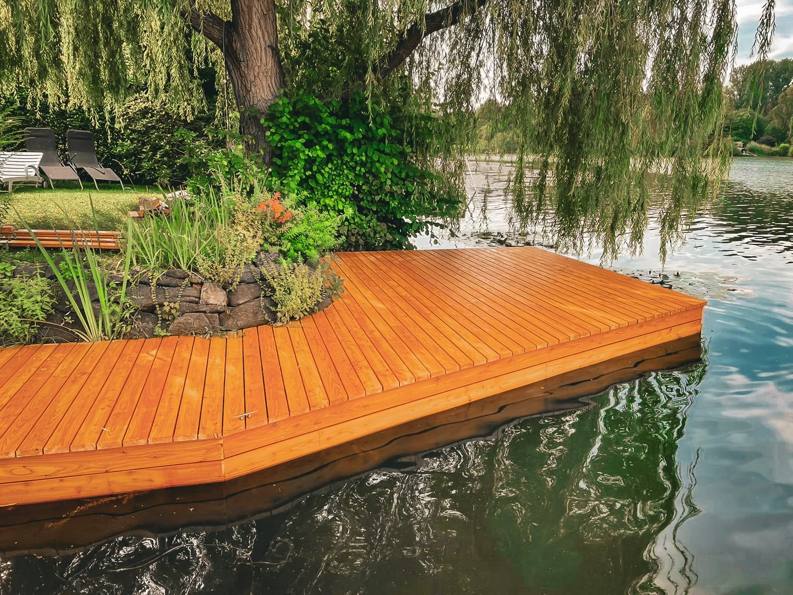 Ein Holzsteg am Wasser, umgeben von Bäumen und Pflanzen, mit Liegestühlen im Hintergrund.