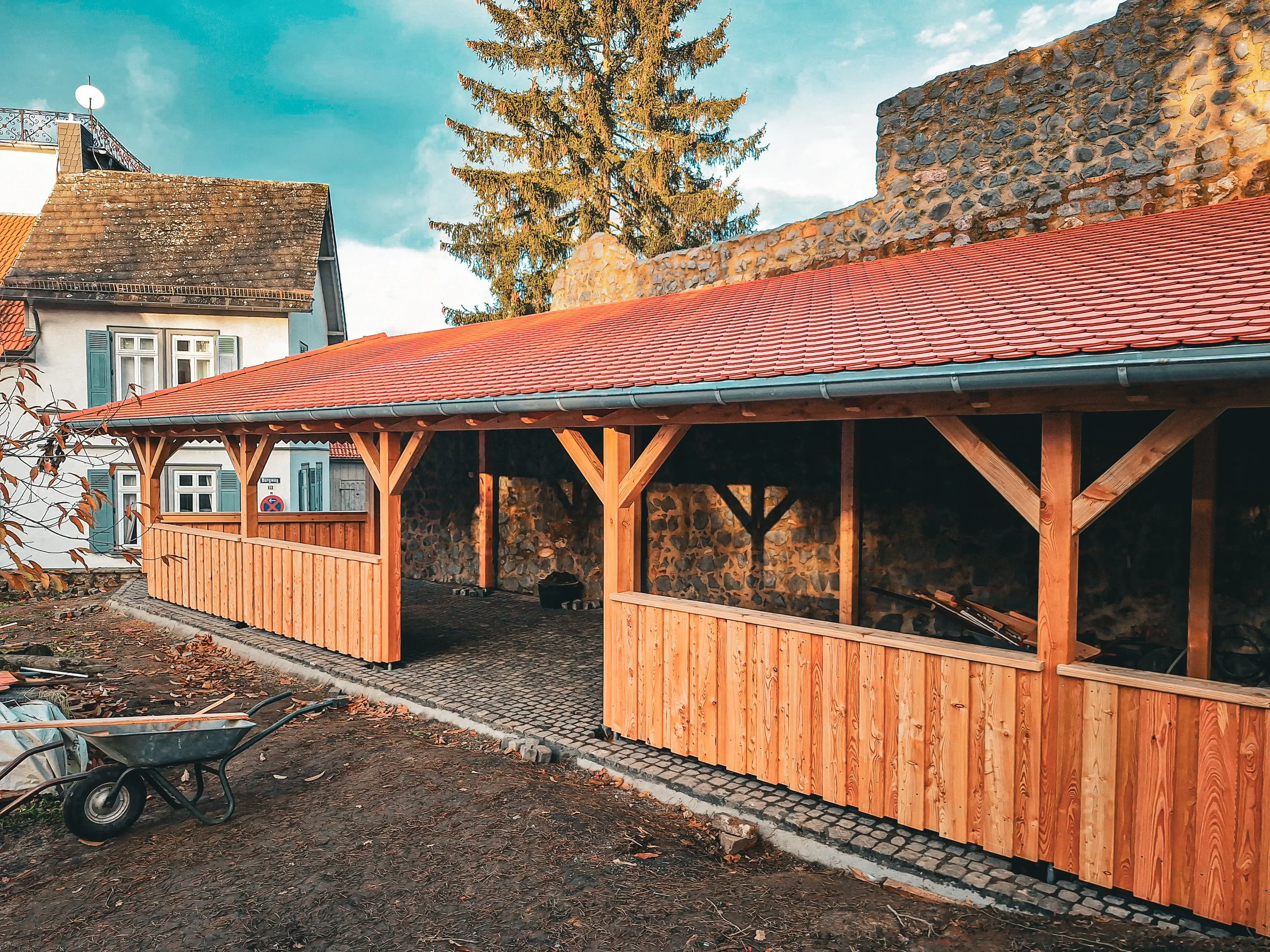 Ein Haus mit weißen Wänden, blauen Fensterläden, einem roten Ziegeldach und einer hohen Tanne im Hintergrund. Im Vordergrund steht ein Unterstand aus Holz mit einem roten Ziegeldach, darunter eine gepflasterte Fläche. Auf der linken Seite befindet sich eine Schubkarre.