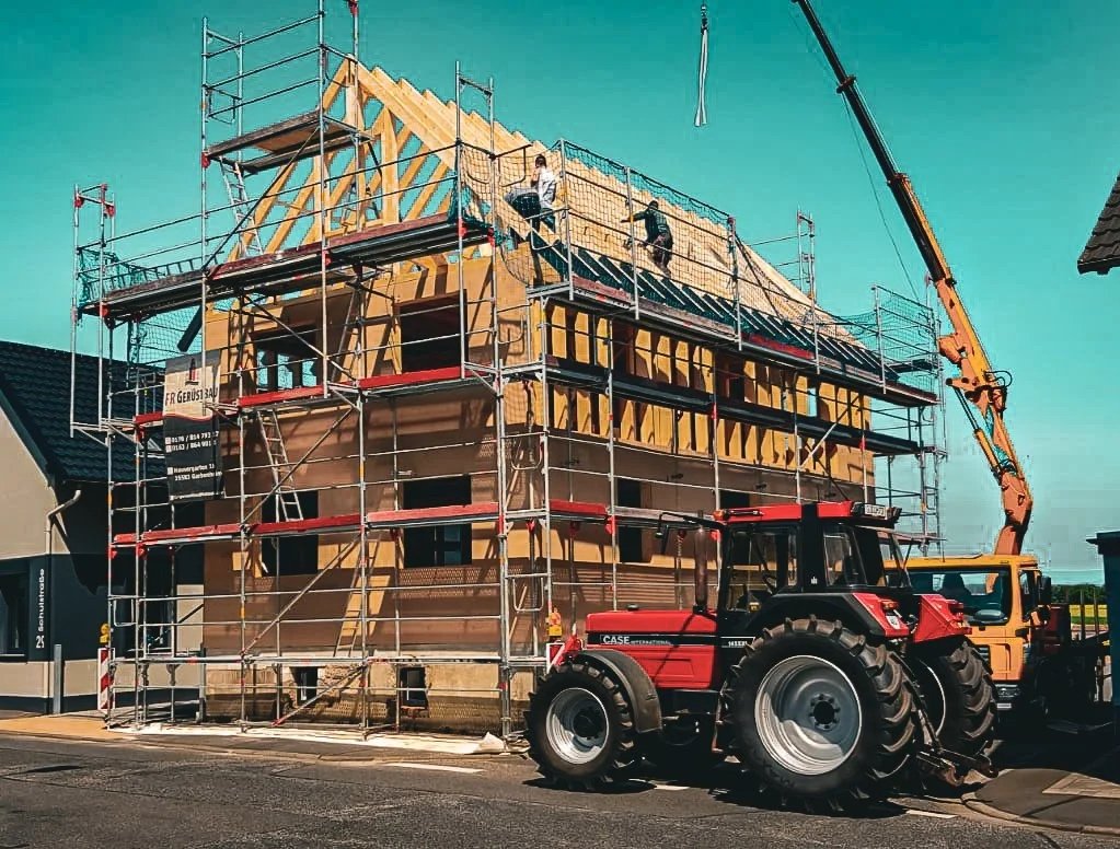 Ein Haus im Bau mit Holzrahmen, umgeben von Gerüsten und einem Kranwagen, während Bauarbeiter die Dacharbeiten durchführen.