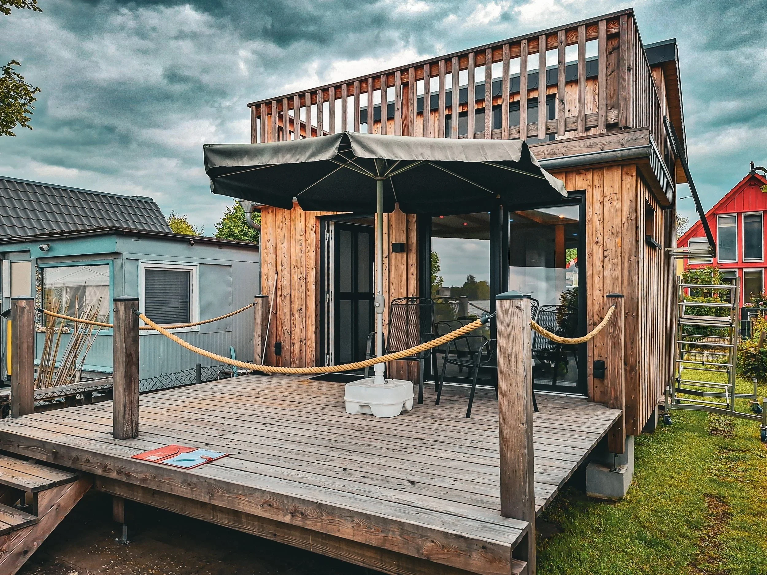 Ein mehrstöckiges Haus mit Holzfassade, Balkon und Terrasse mit einem großen Sonnenschirm. Umgeben von einem Garten und unter einem bewölkten Himmel.