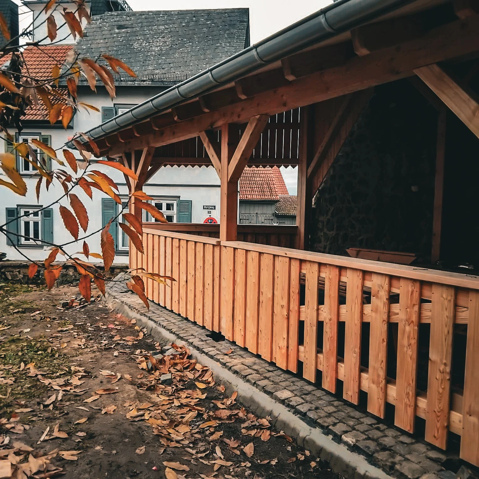Holzveranda mit Dachüberhang und Zaun vor einer weißen Hauswand, im Vordergrund Blätter auf dem Boden, herbstliche Atmosphäre.