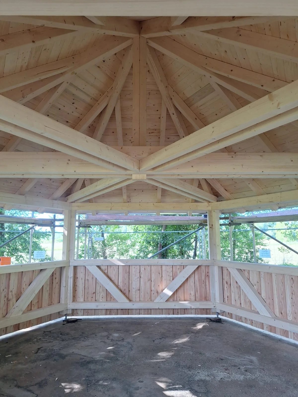 Innenaufnahme einer Holzbaukonstruktion in einem Pavillon oder einer Hütte, mit sichtbarem Dachstuhl und umlaufender Brüstung, umgeben von Bäumen.