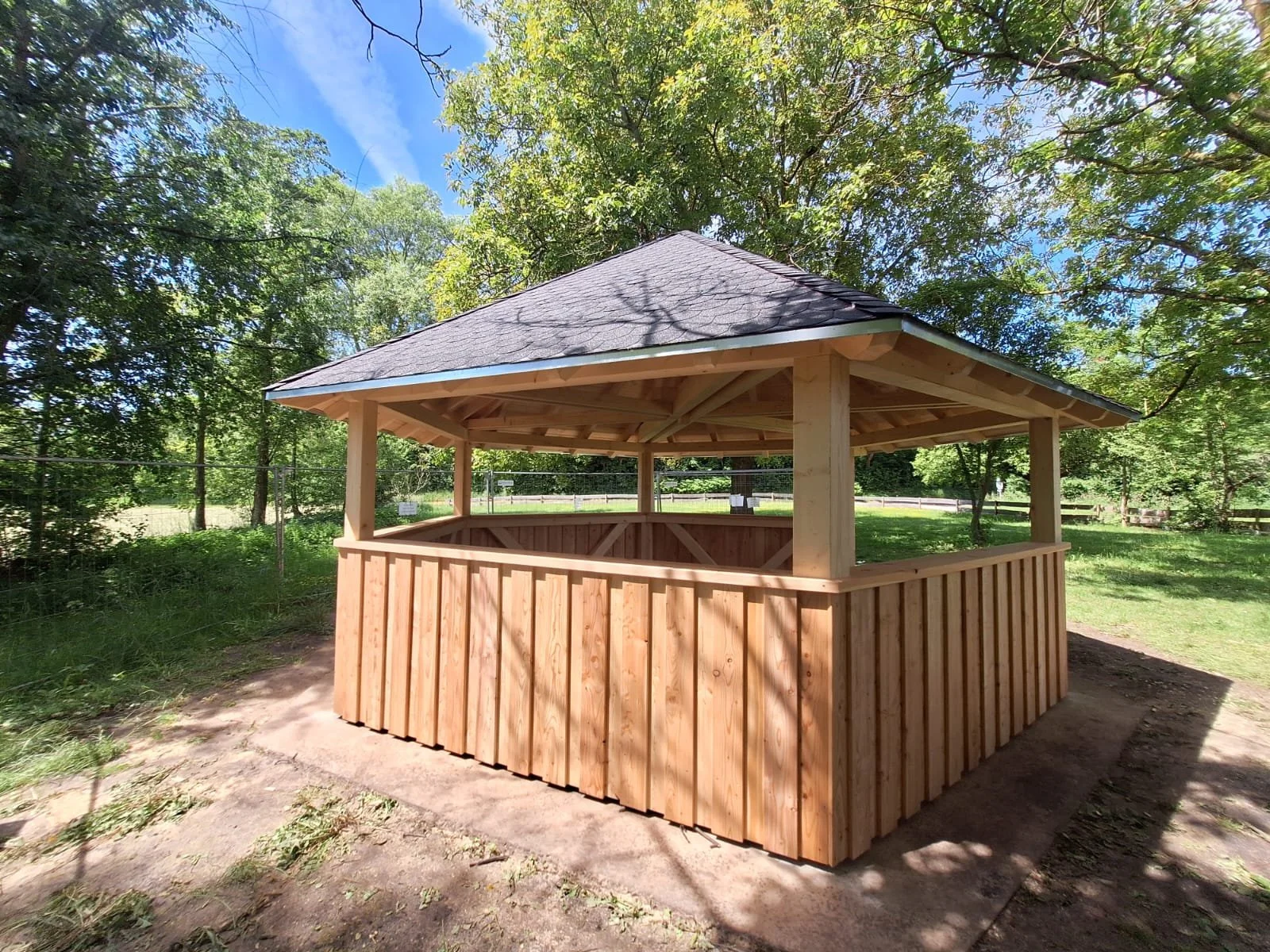 Ein Holzpavillon in einem Park mit umgebenen Bäumen und Zaun, sonnig bei blauem Himmel.