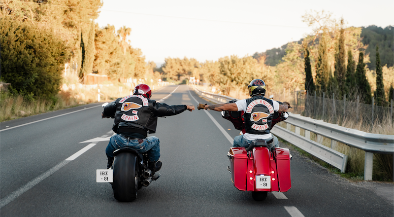 Dos motociclistas con chaquetas de motorista con parches de España y Los Ángeles, circulando por una carretera de dos carriles, tomados de la mano y extendidos a lo largo de la autopista mientras iban uno al lado del otro al final de la tarde.