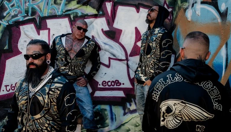 Rockeros con tatuajes y gafas de sol de pie junto a una pared grafitada, vistiendo sudaderas negras con diseños dorados y negros.