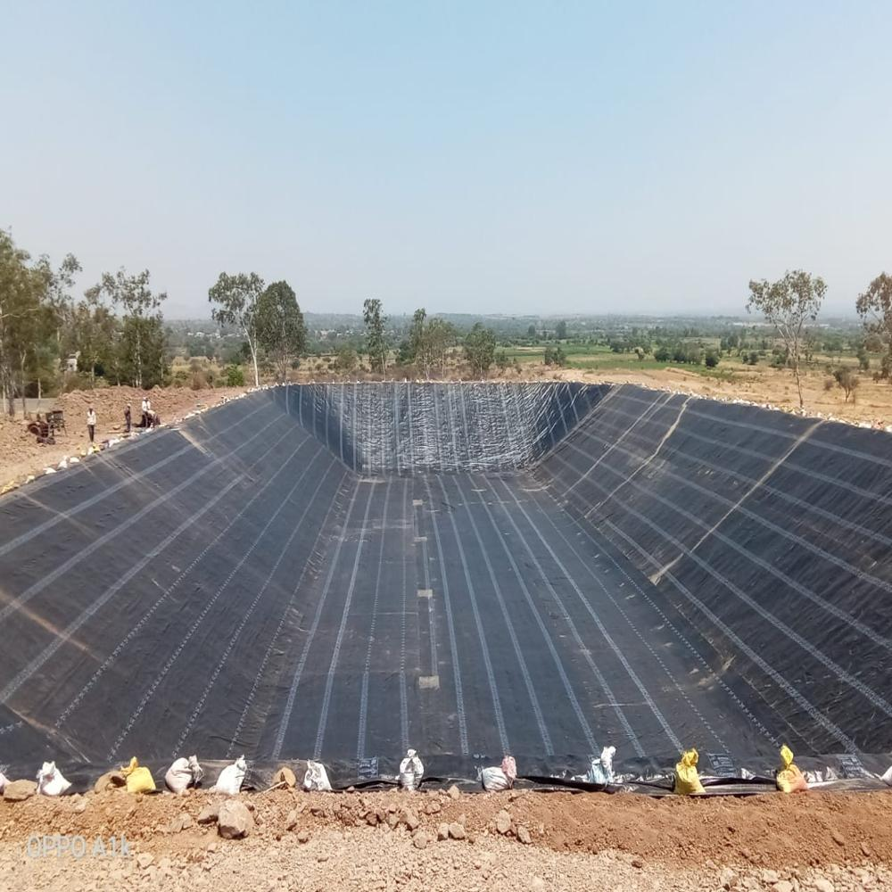 A large rectangular pond with a covering of plastic mesh, situated in an open field with green trees and a small building in the background.