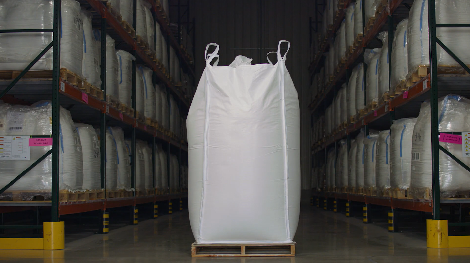A large white industrial bag stands on a pallet in the center of a warehouse aisle, surrounded by shelves filled with similar bags.