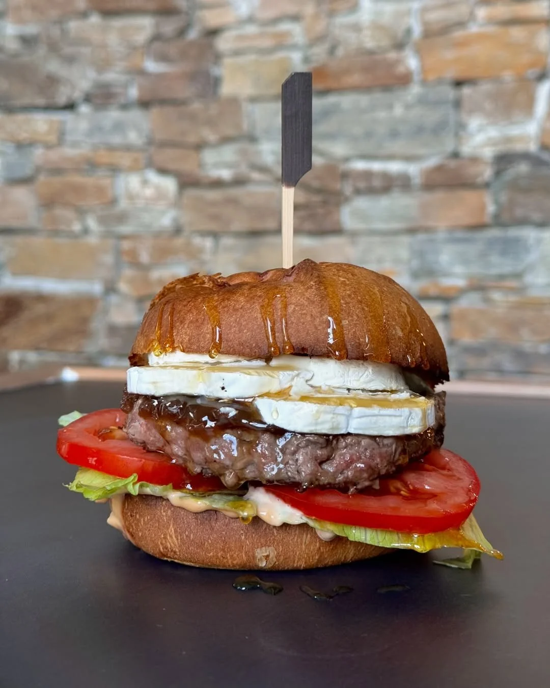 Un burger avec un pain, une tranche de tomate, de la laitue, une galette de viande, des tranches de fromage, un œuf au plat et une sauce, présenté sur une surface noire avec un mur en briques en arrière-plan.