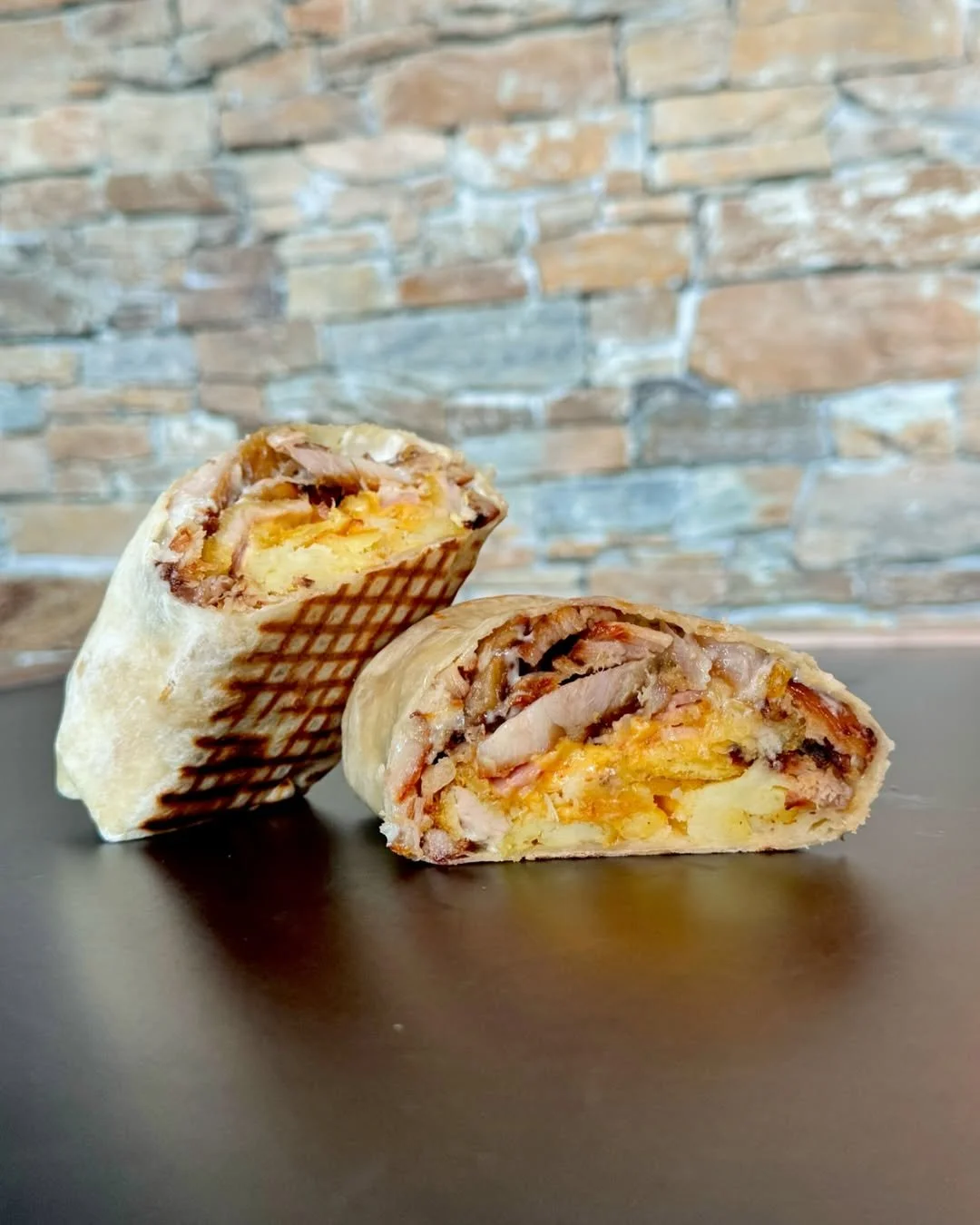 Un wrap coupé en deux contenant du poulet, du fromage et des pommes de terre, posé sur une surface noire, avec un mur en pierre en arrière-plan.