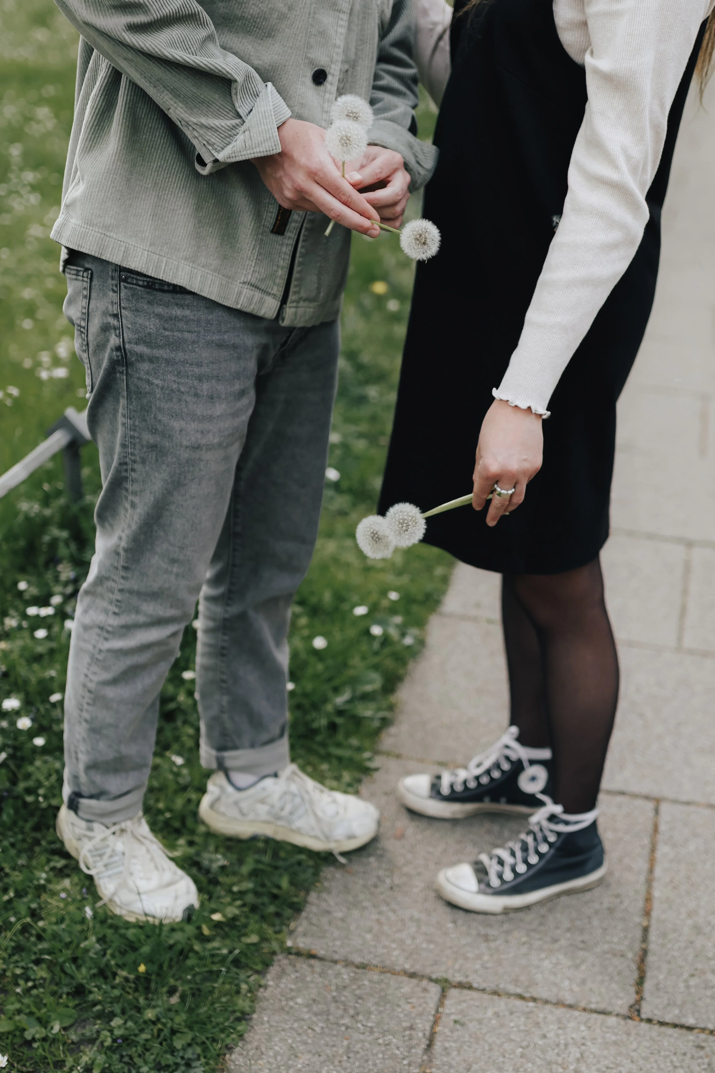 Zwei Menschen stehen auf einem Gehweg im Freien, beide halten Löwenzahnblumen. Eine Person trägt eine graue Jacke und Jeans, die andere trägt einen schwarzen Rock, Tights und Sneakers.