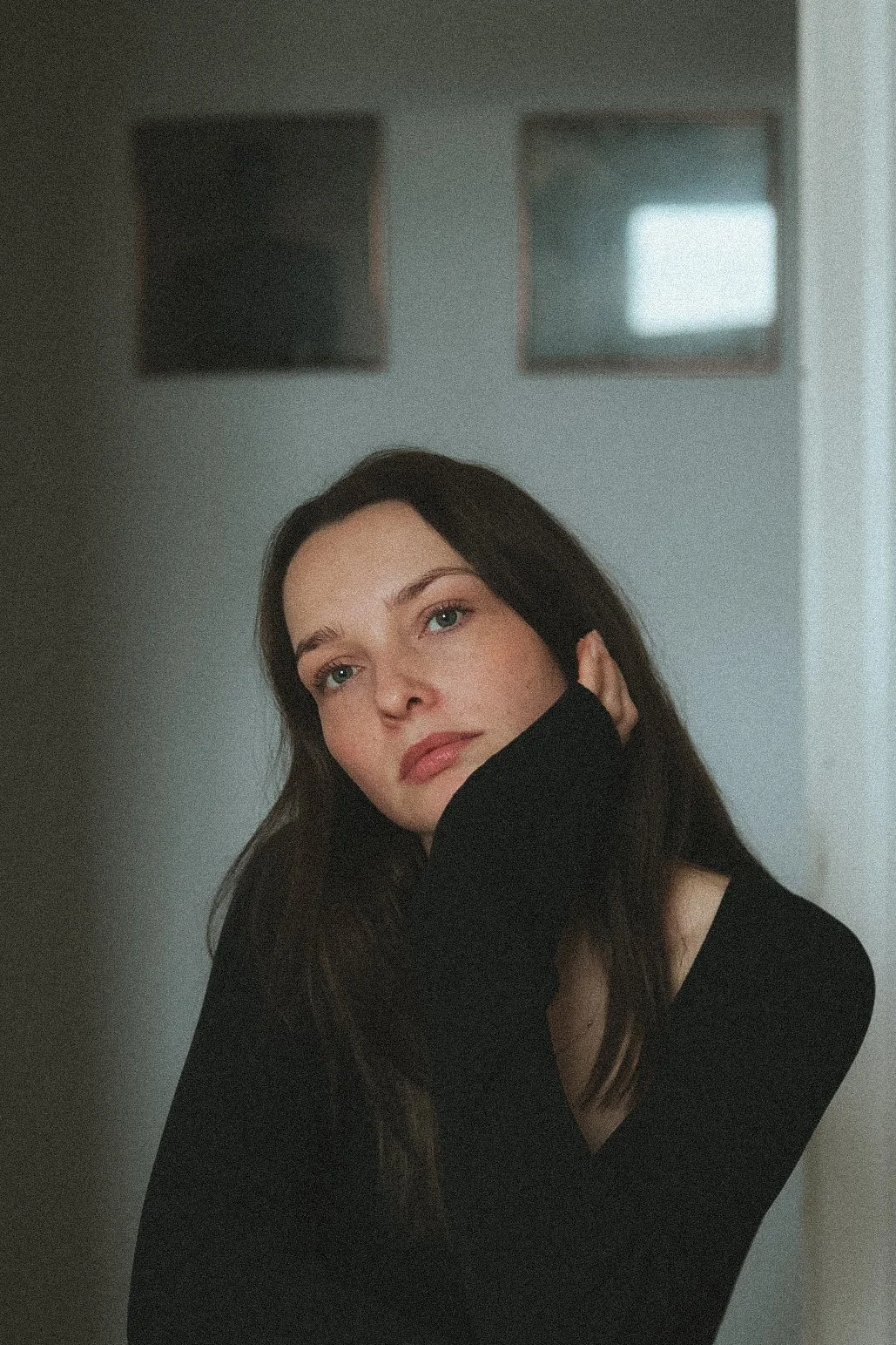 Porträt einer jungen Frau mit langen braunen Haaren, schwarzen Pullover, blickt nach oben, in einem Raum mit zweifach quadratischen Fenster im Hintergrund.