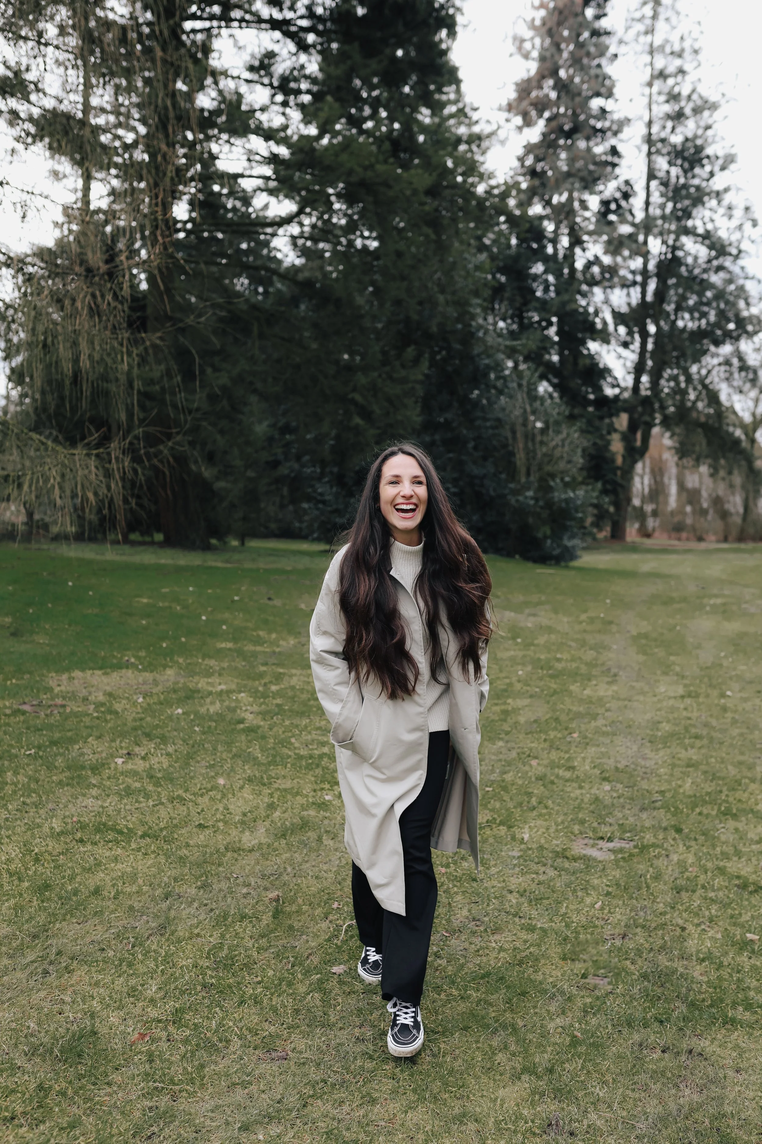 Junge Frau mit langen, lockigen braunen Haaren, lachend im Park, trägt eine beige Steppjacke, schwarze Hose und Vans Sneakers.