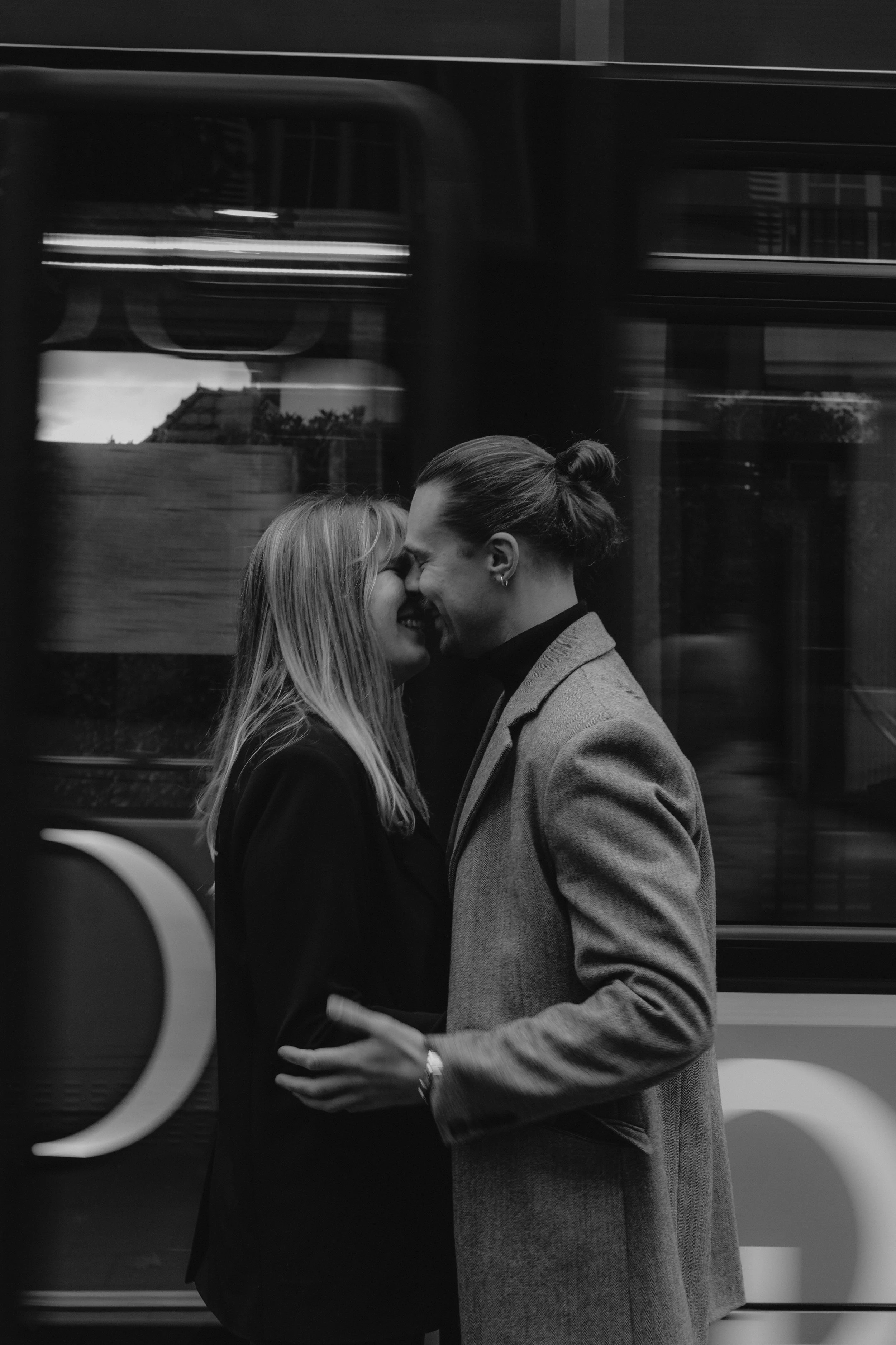 Ein Paar, ein Mann und eine Frau, küsst sich in einer öffentlichen Umgebung, vermutlich in einer Stadt, während eine Straßenbahn im Hintergrund vorbeifährt.