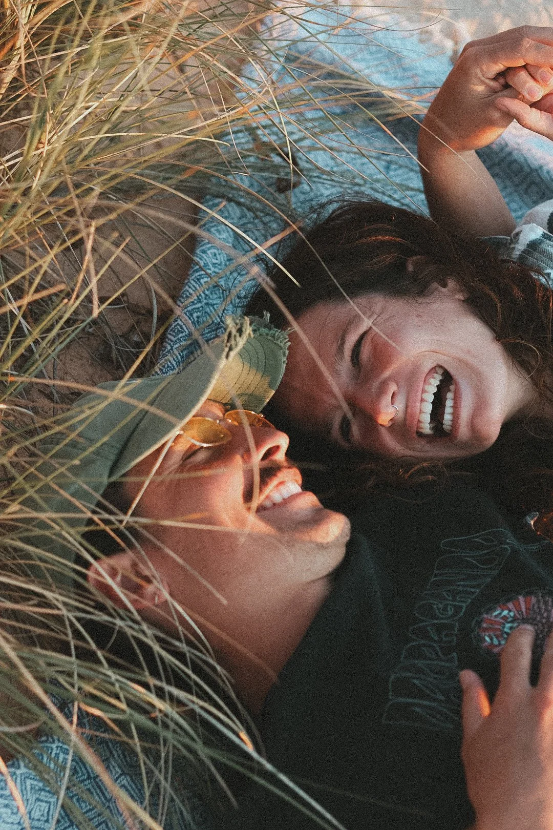 Zwei Menschen liegen lachend im Sand zwischen Strandgras, eine trägt Sonnenbrille und das andere trägt einen Nagelring.