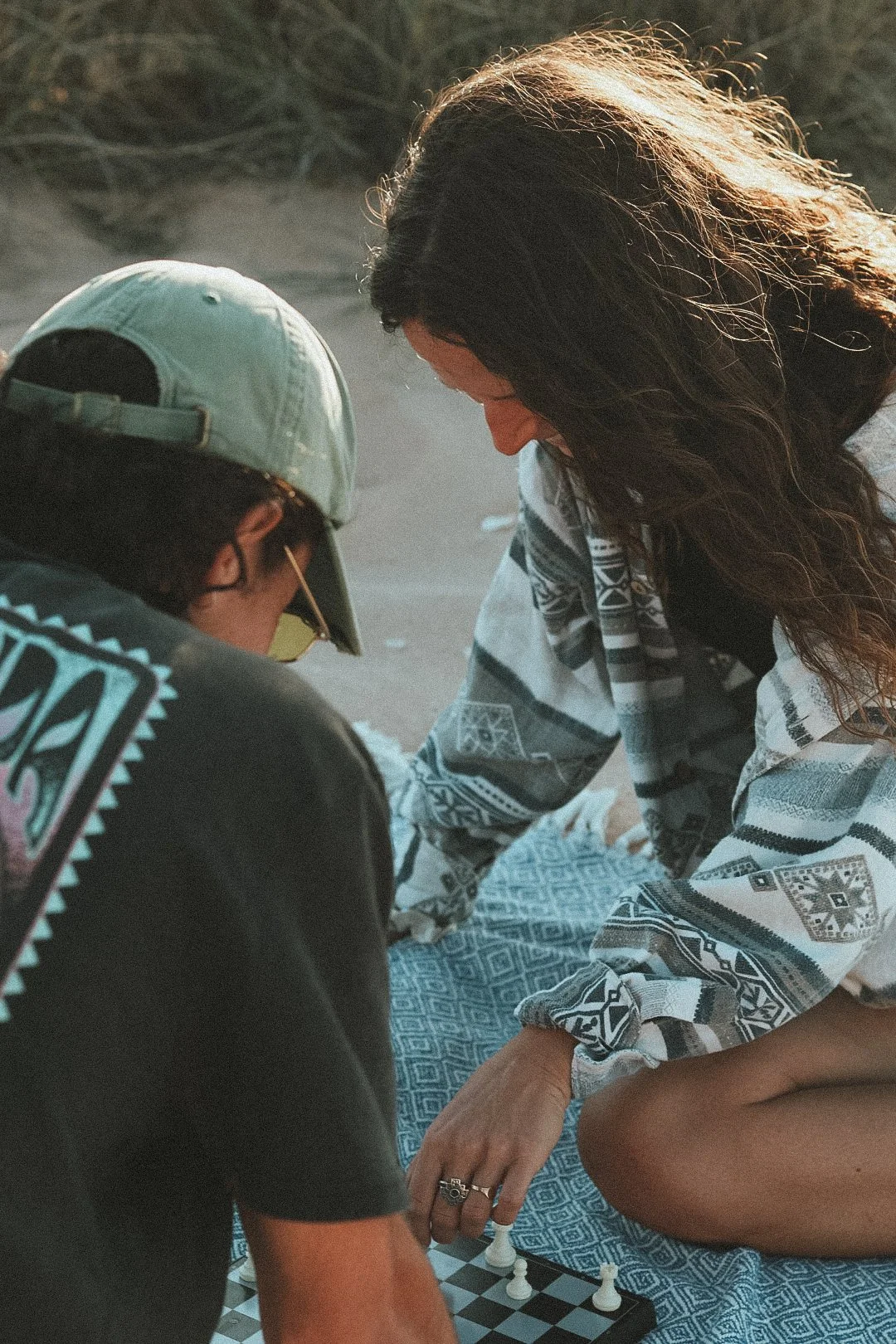 Zwei Menschen spielen Schach im Freien auf einer Decke, eine Frau und ein junger Mann, der ein Cap trägt.