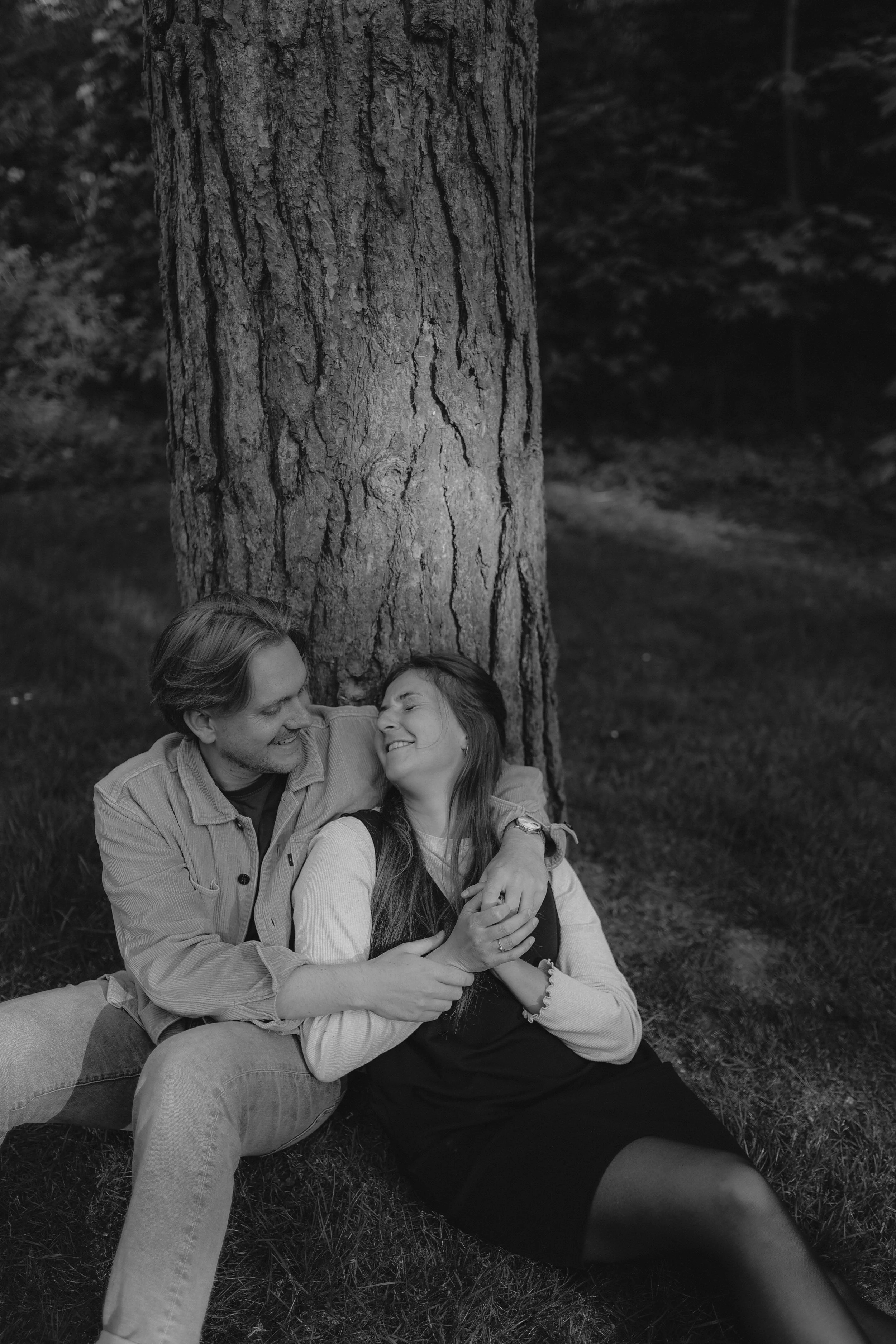 Ein Mann und eine Frau sitzen lachend und umarmt an einem Baum im Freien.