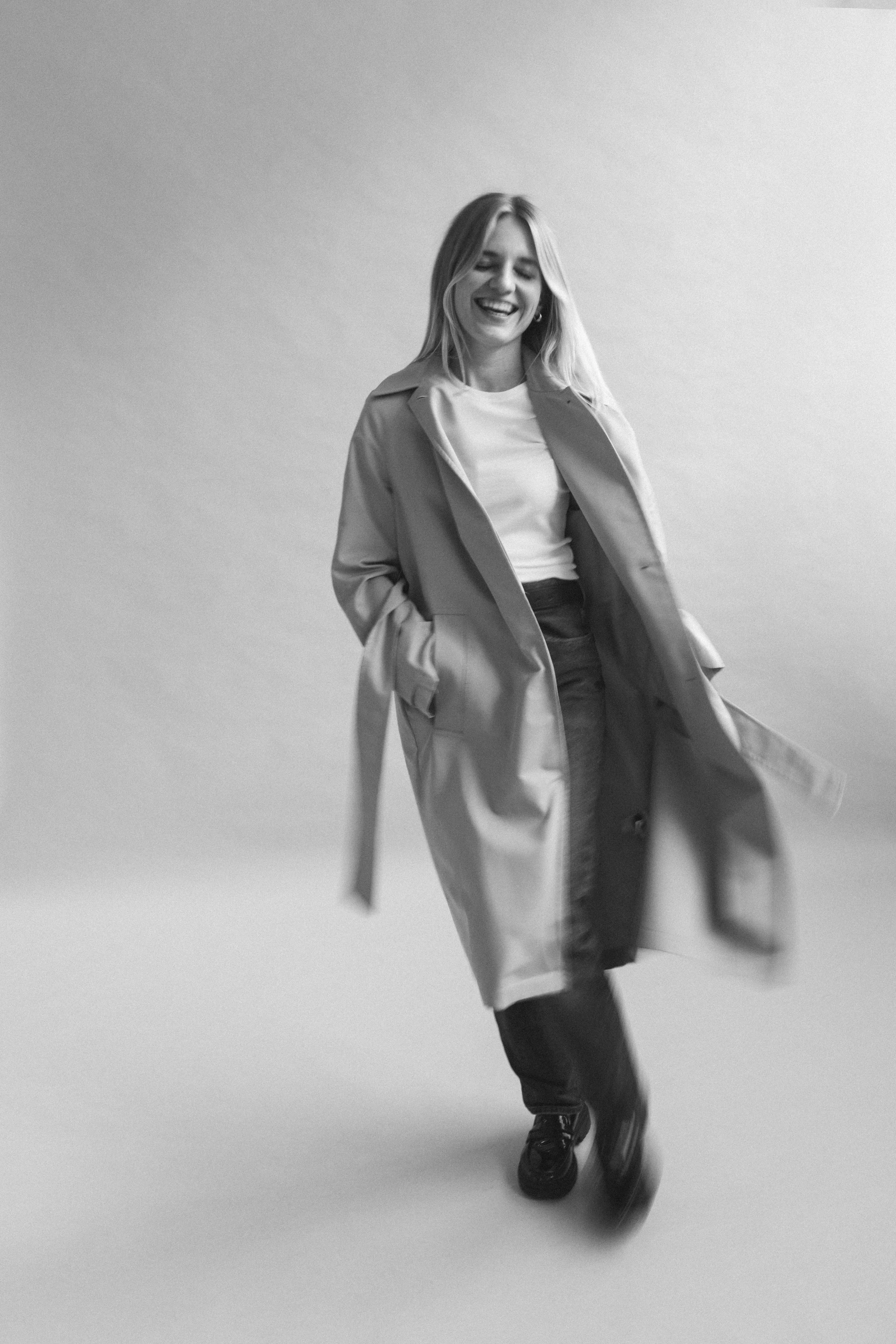 Eine lachende junge Frau mit langem Haar trägt einen Trenchcoat, weiße T-Shirt, dunkle Hosen und Schuhe, sie läuft lachend auf einer neutralen, hellen Hintergrund.