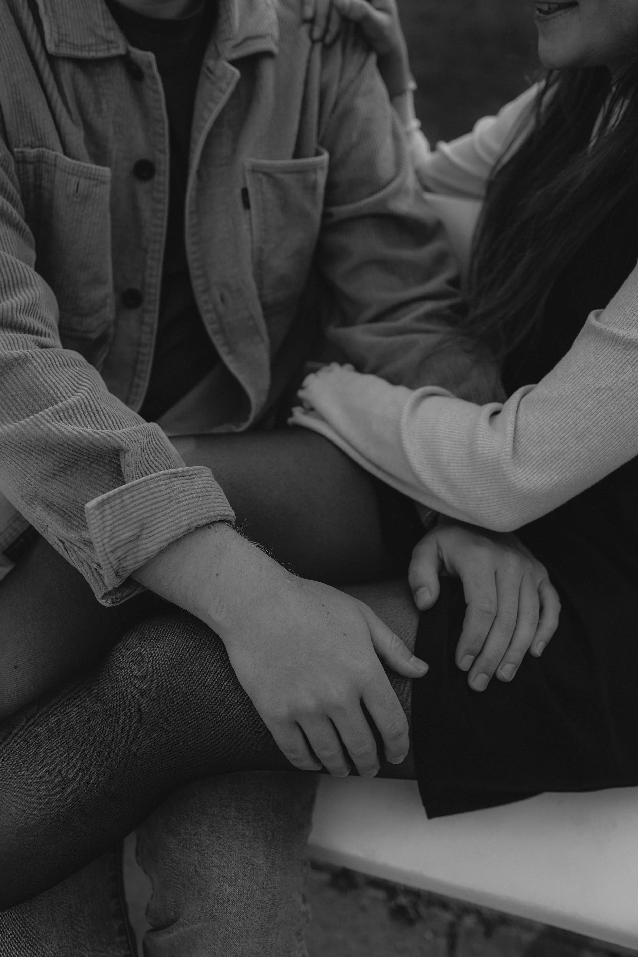 Zwei Menschen sitzen eng beieinander, einer berührt den anderen an der Schulter, die Hände liegen auf den Beinen, eine Person trägt einen Jeansjacke, die andere eine helle Jacke.