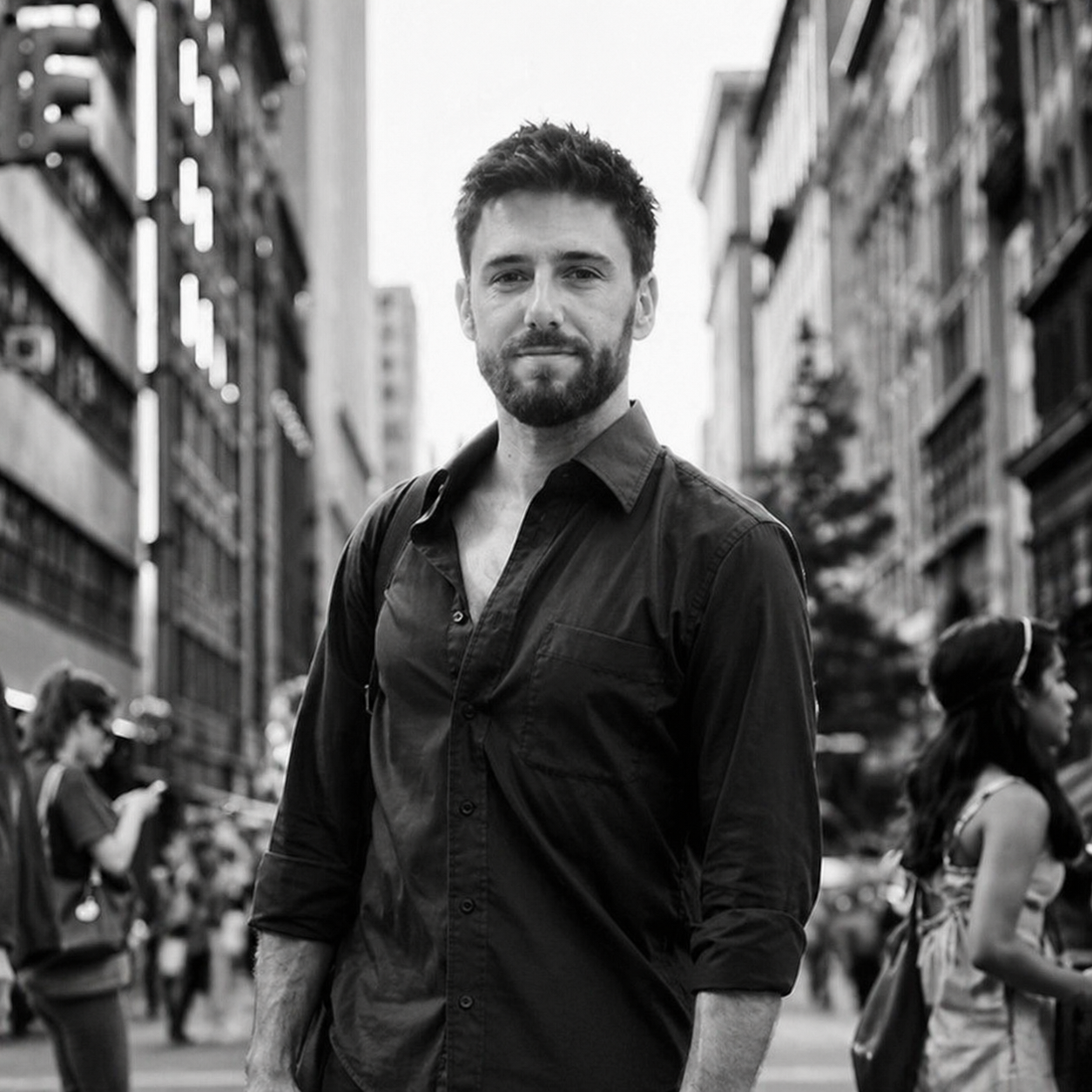 A black and white photo of a young man with a beard and short hair, wearing a button-up shirt, standing on a city street with tall buildings and other people in the background.