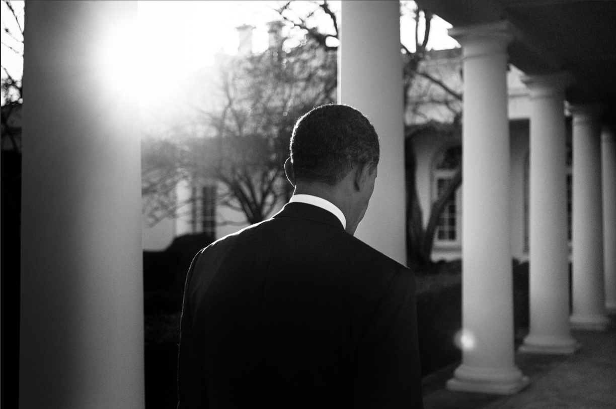 A man in a suit standing outdoors between white columns during sunset, viewed from behind in black and white.