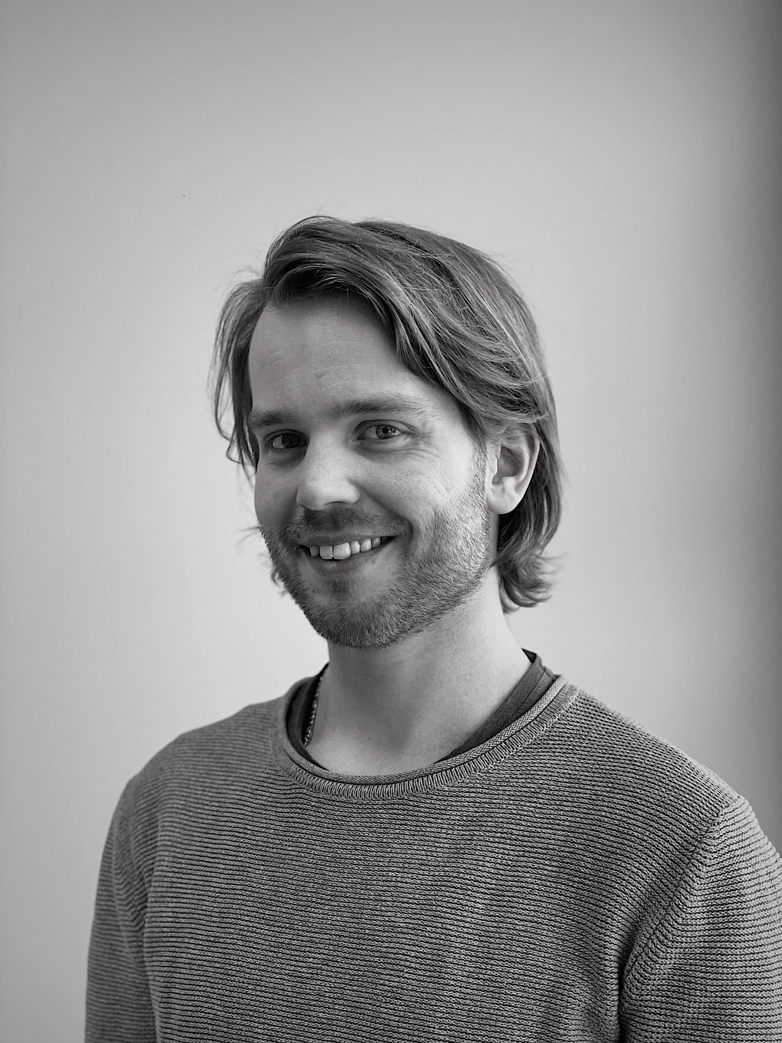 Portrett av en mann med skulderlangt hår og skjegg, smilende mot kamera, iført en stripet genser, mot en enkel lys bakgrunn.